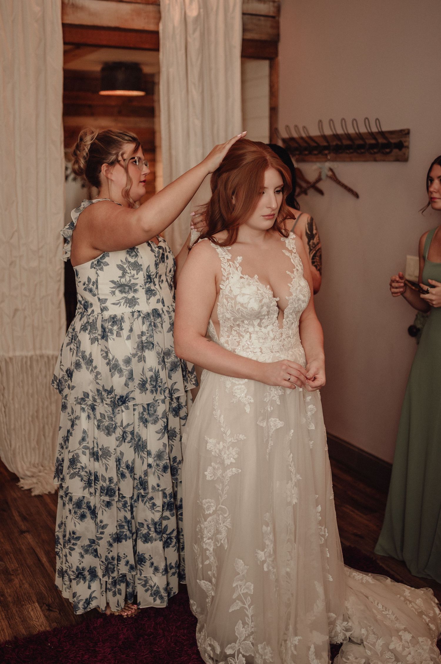A bride in a white lace gown gets help with her dress in a rustic indoor setting.