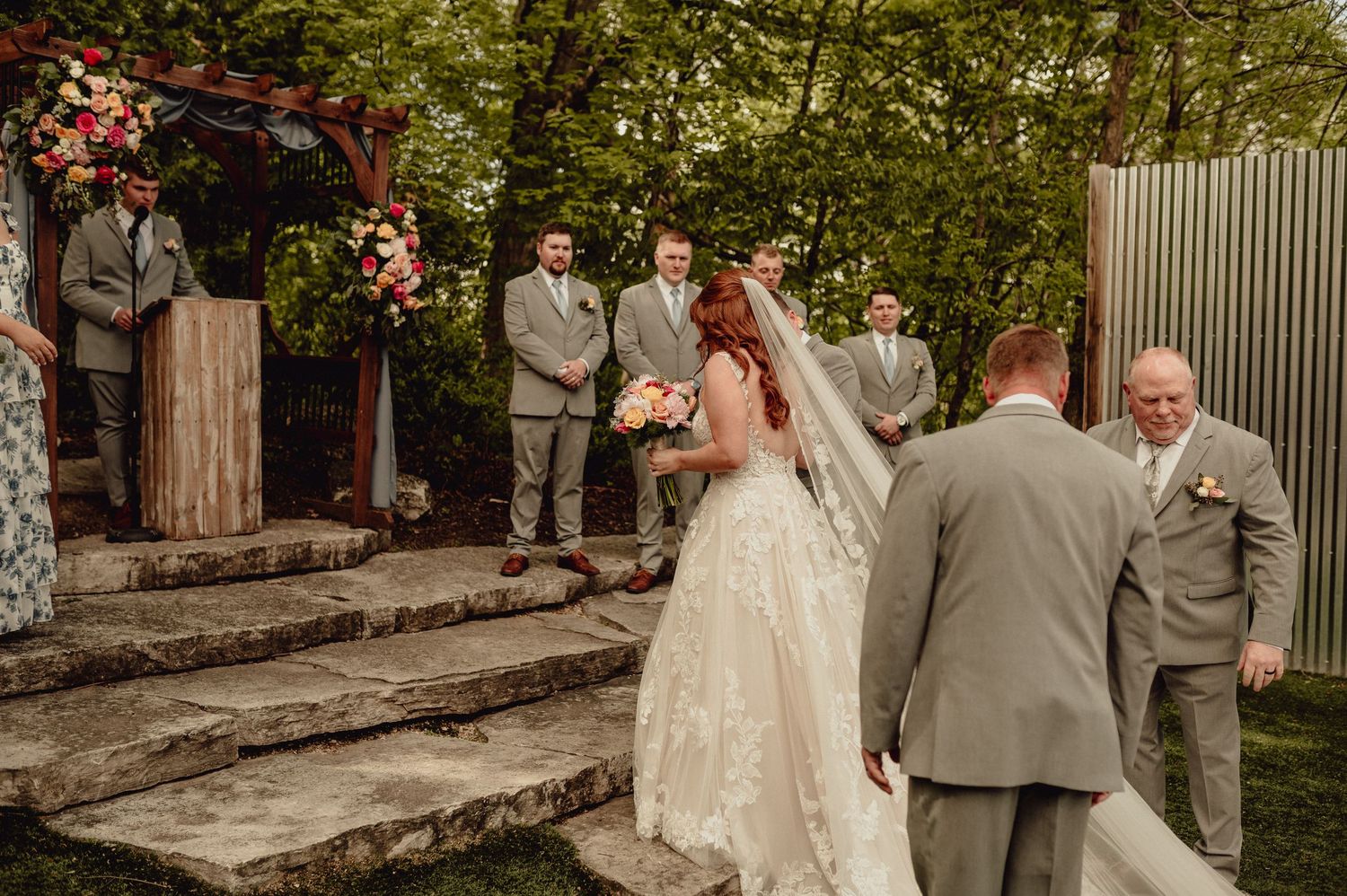 Wedding party stands on stone steps surrounded by lush greenery and pink flowers in an outdoor garden setting.