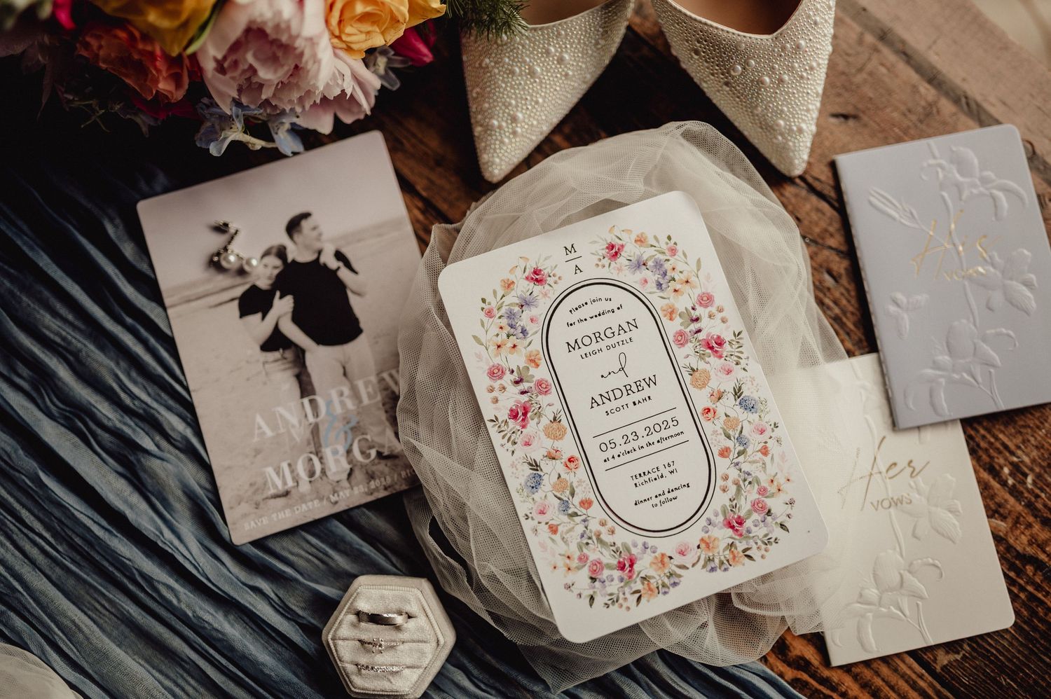 Wedding invitations with floral designs and calligraphy laid out on a dark wooden surface with dried flowers.