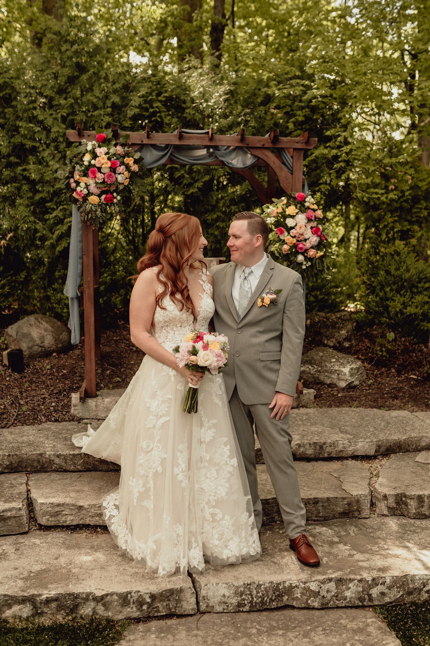 A couple in wedding attire sits together on stone steps surrounded by lush greenery and floral arrangements.