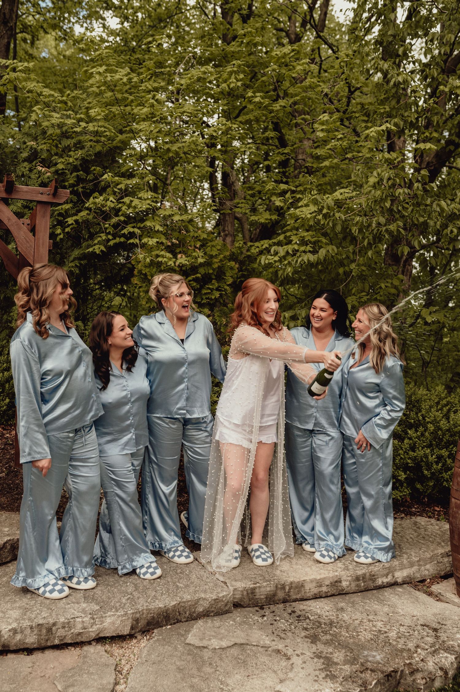 Bridesmaids in coordinated blue dresses share a candid laughing moment while sitting on a concrete wall outdoors.