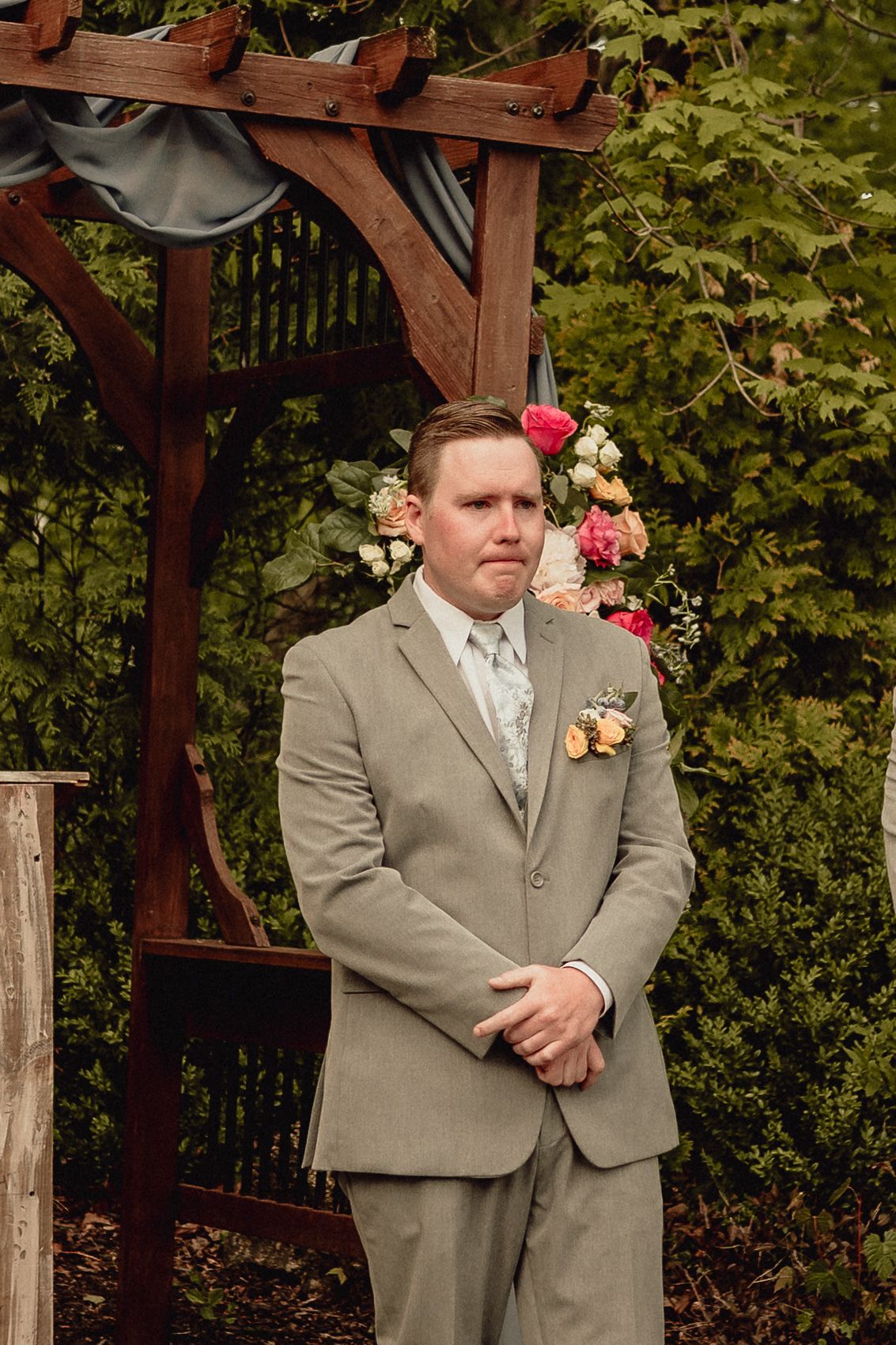 Wedding party members in gray suits stand in a row under a wooden pergola with pink flowers and greenery.