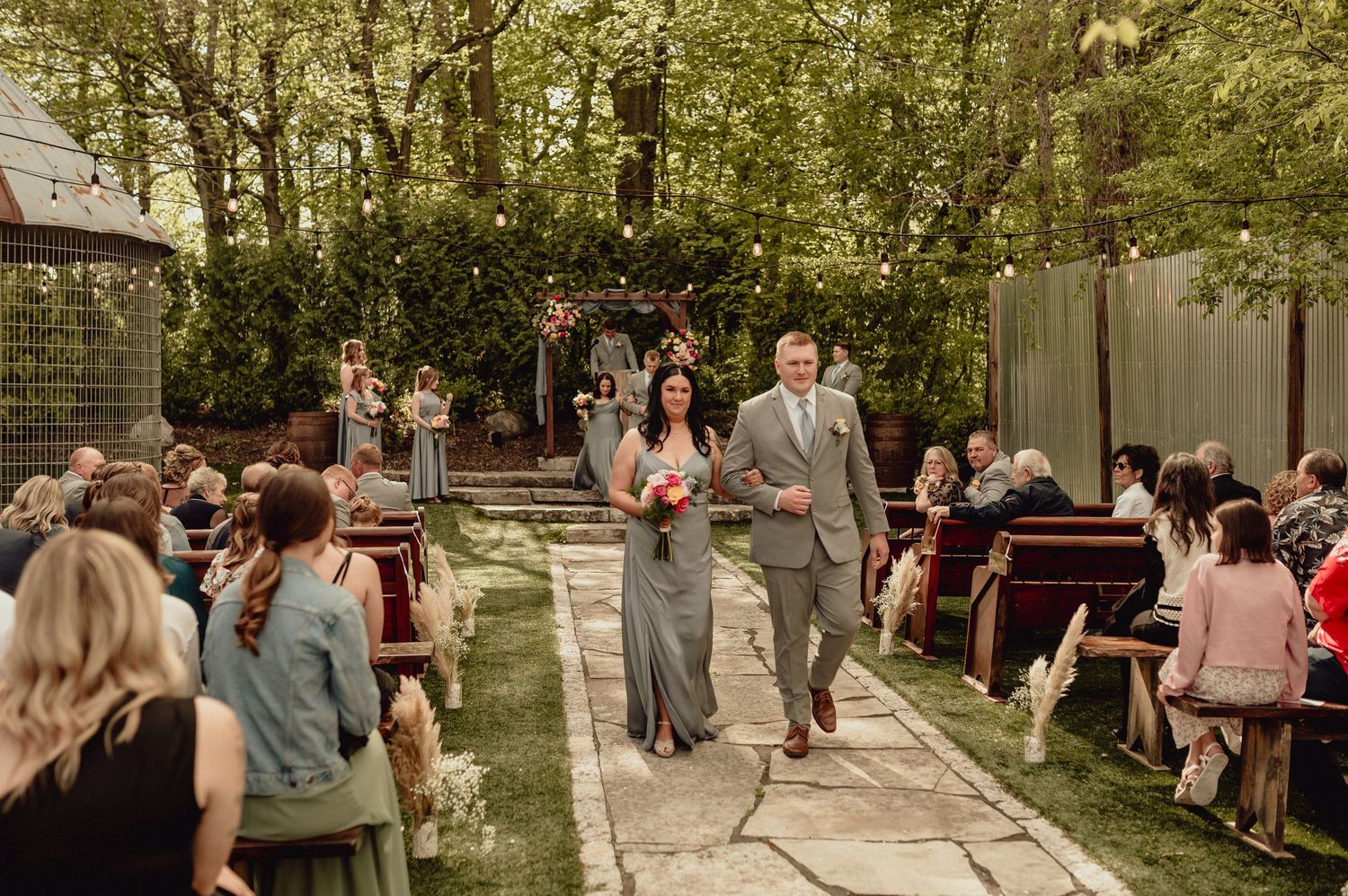 Outdoor garden wedding ceremony with guests seated on wooden benches along a stone pathway surrounded by trees.