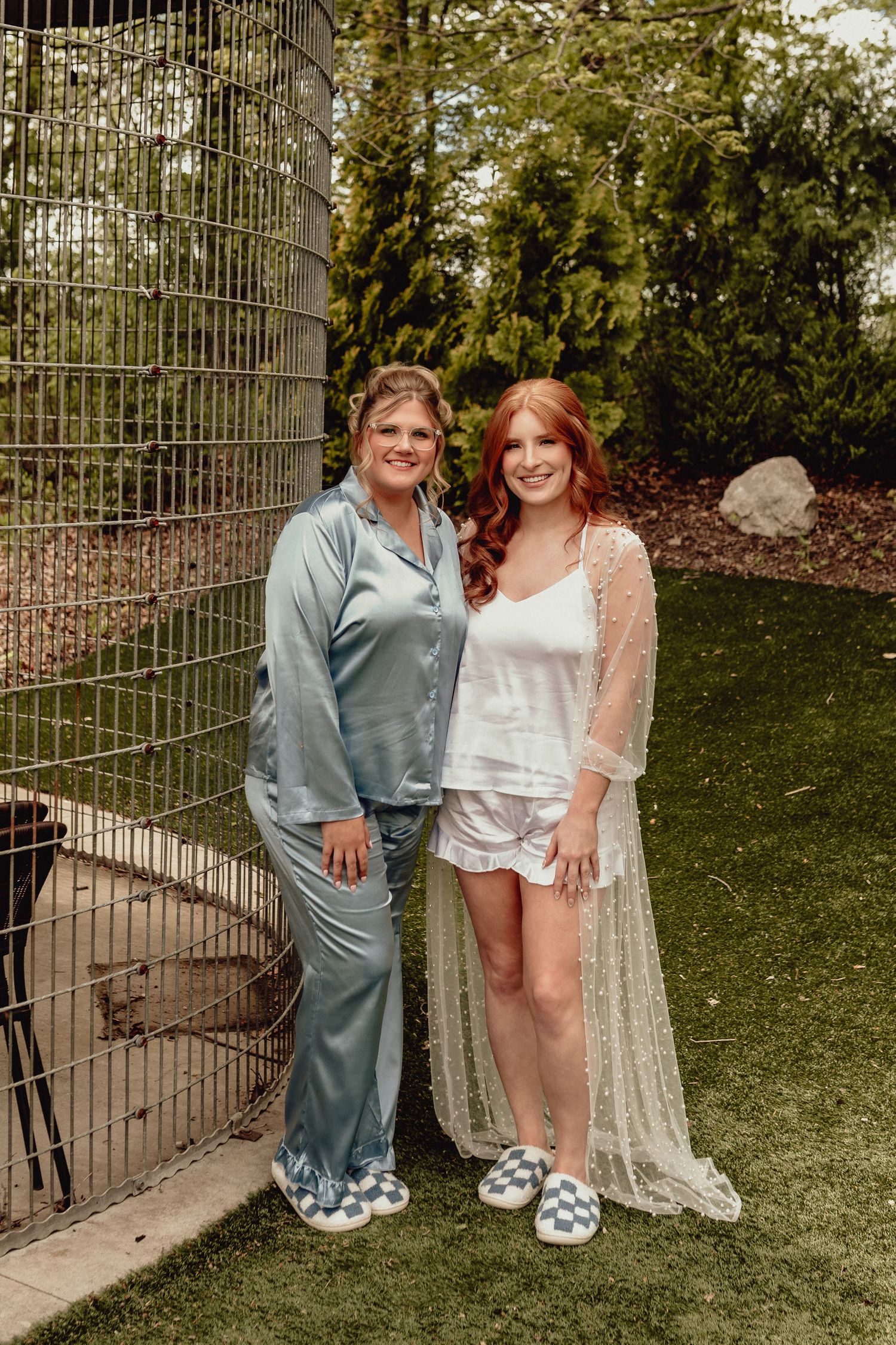A couple poses together outdoors by a metal gate, wearing casual attire and cozy slippers.