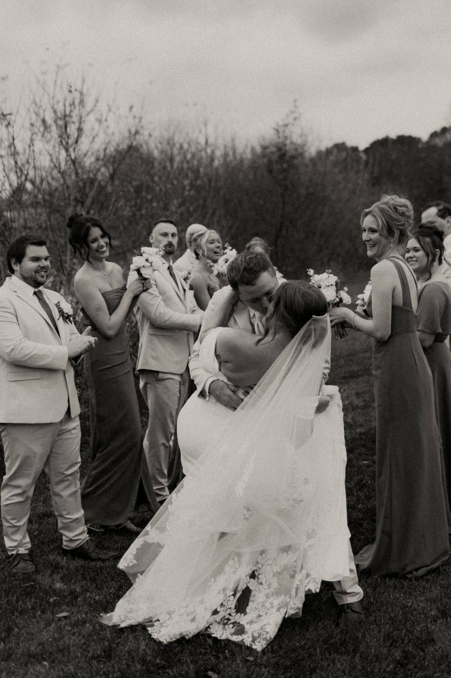 Wedding party members help adjust a bride's flowing white veil outdoors in a series of black and white photographs.
