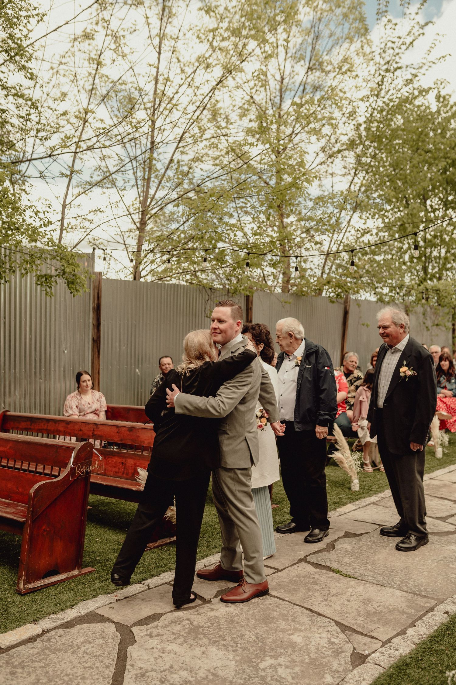 Group of wedding guests perform a synchronized dance line outdoors near a red piano and wooden fence.