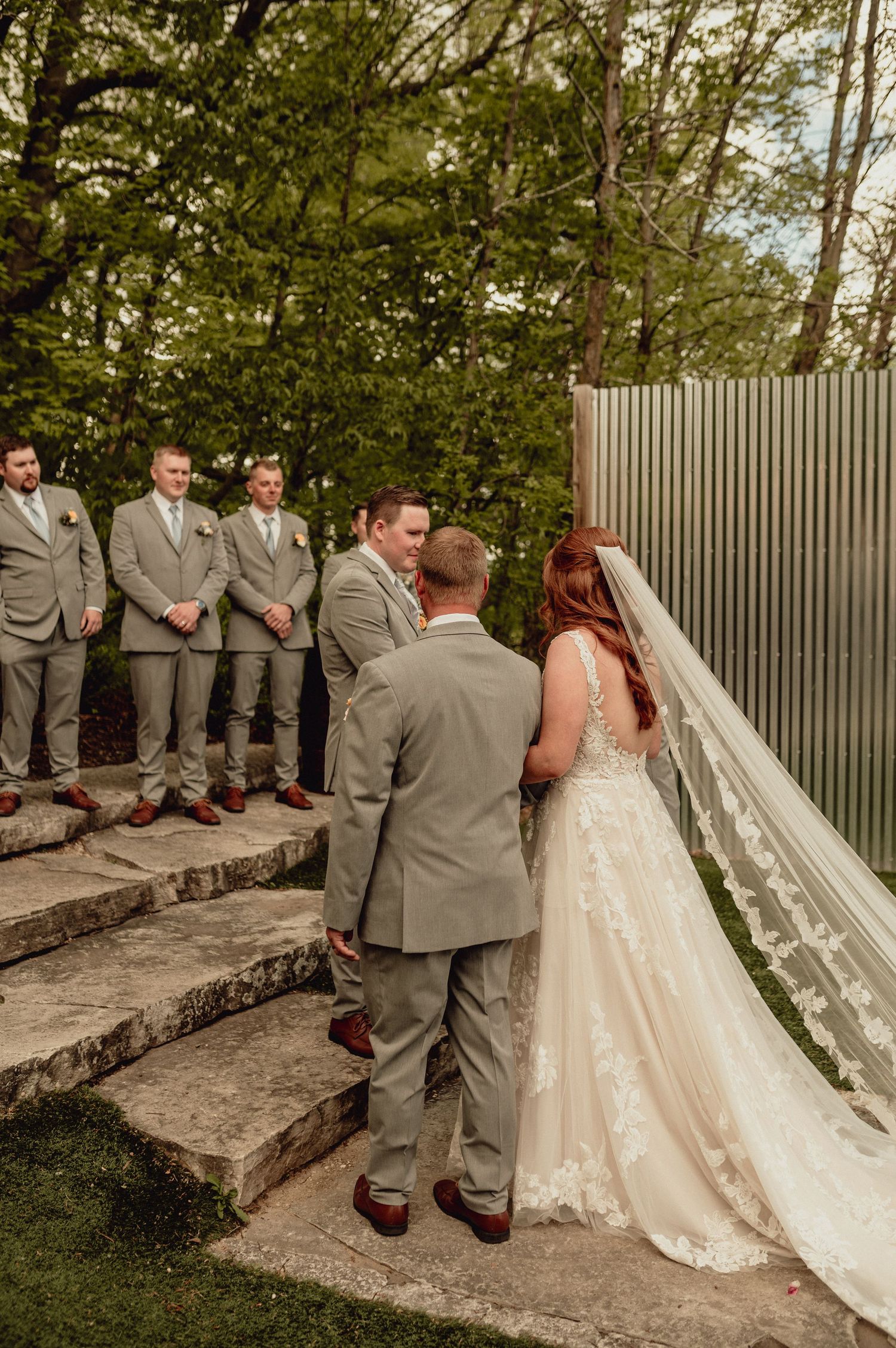 Wedding party in gray suits and white dress pose on stone steps in an outdoor garden setting.