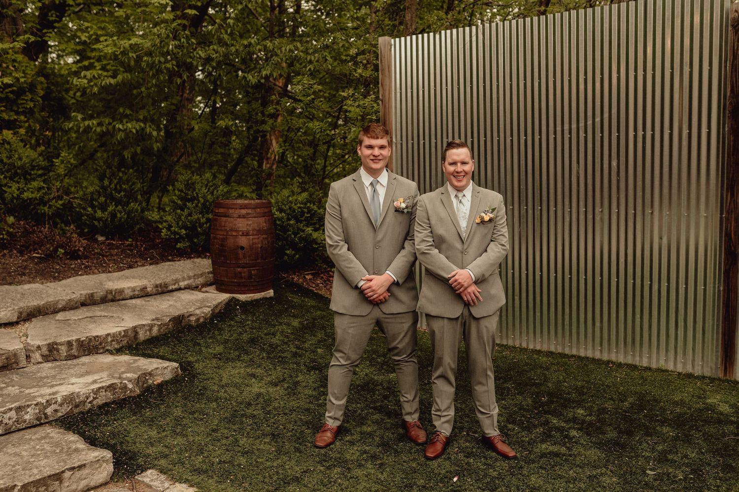 Two people in gray suits sit on stone steps in a garden setting with a metal fence in the background.