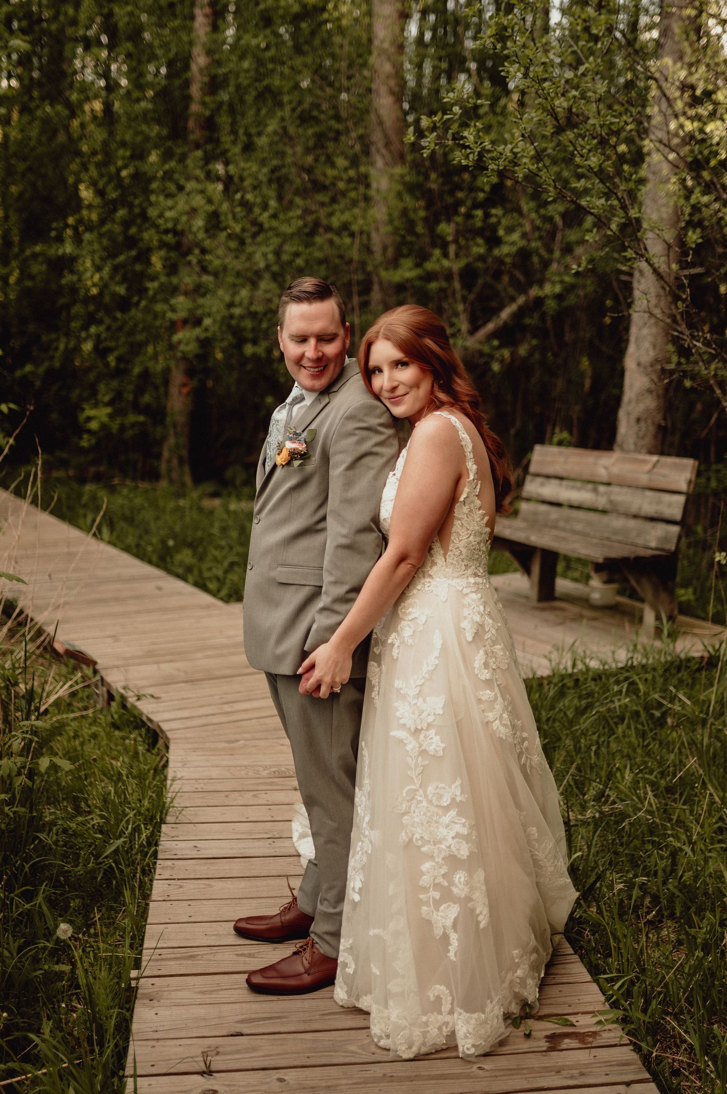A couple in wedding attire poses together on a wooden boardwalk surrounded by lush green foliage.