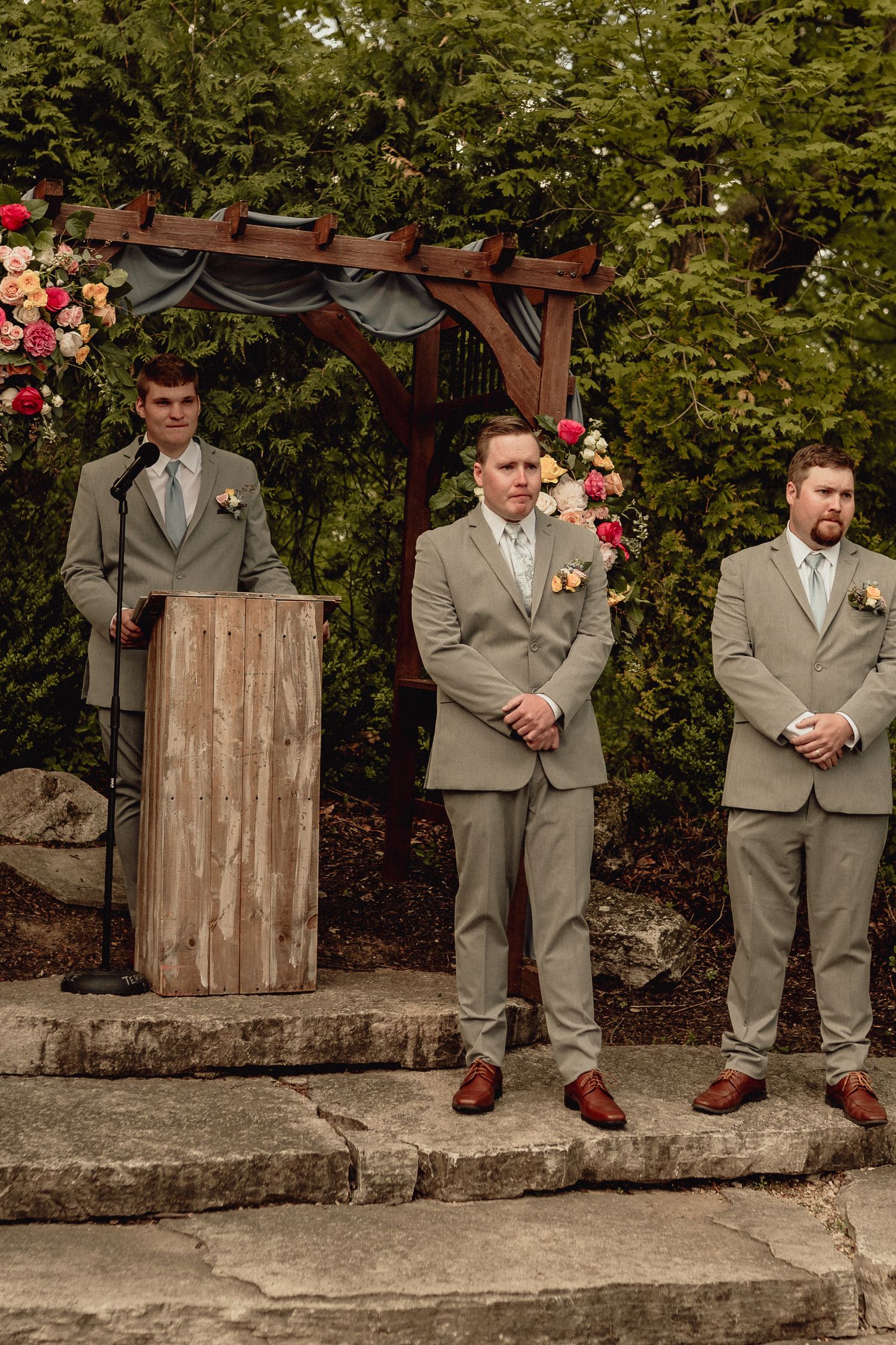 Three groomsmen in gray suits lean against a rustic stone wall with red floral decorations in an outdoor garden setting.