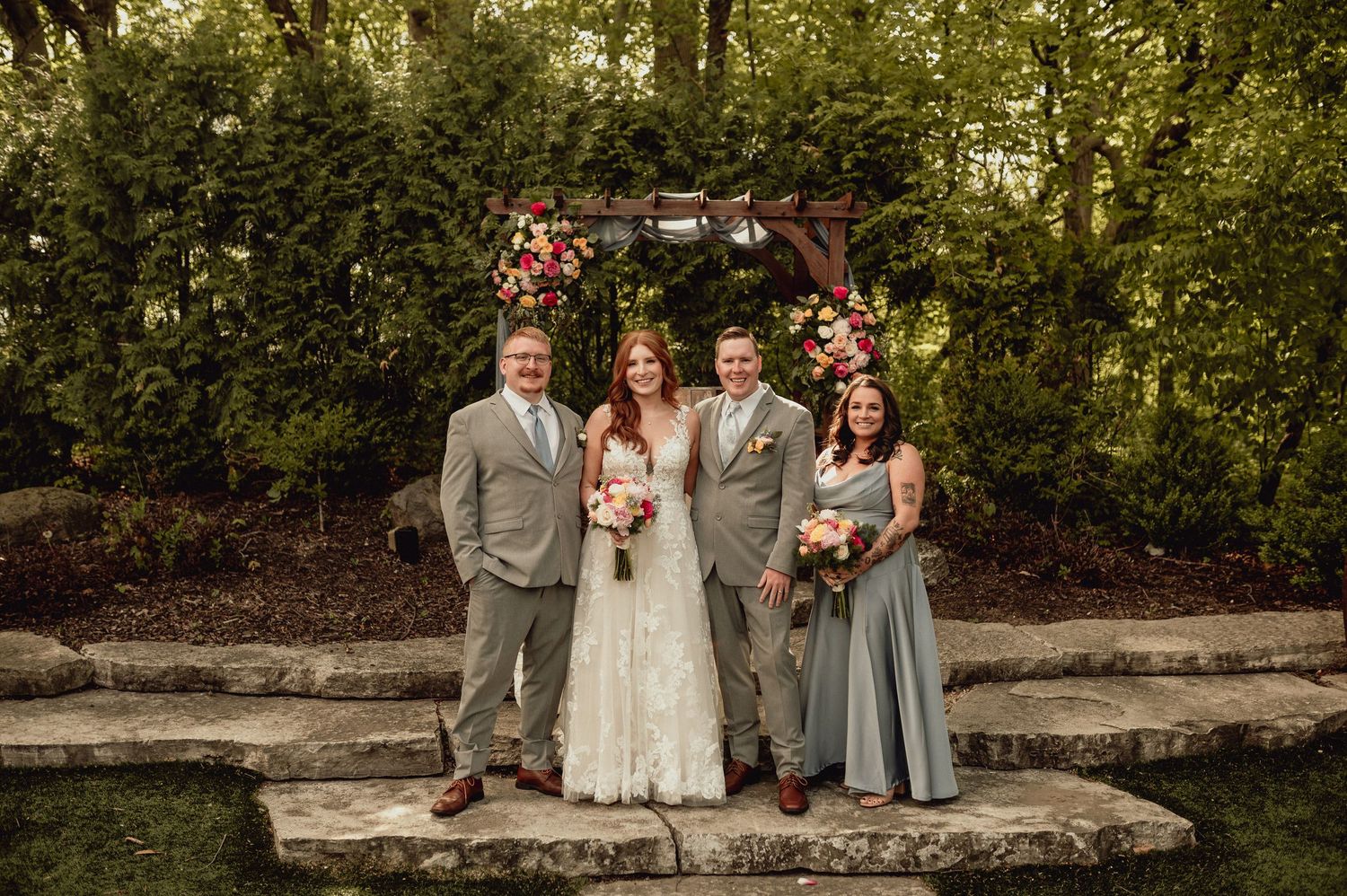Wedding party poses together on stone wall in outdoor garden setting with lush greenery in background.