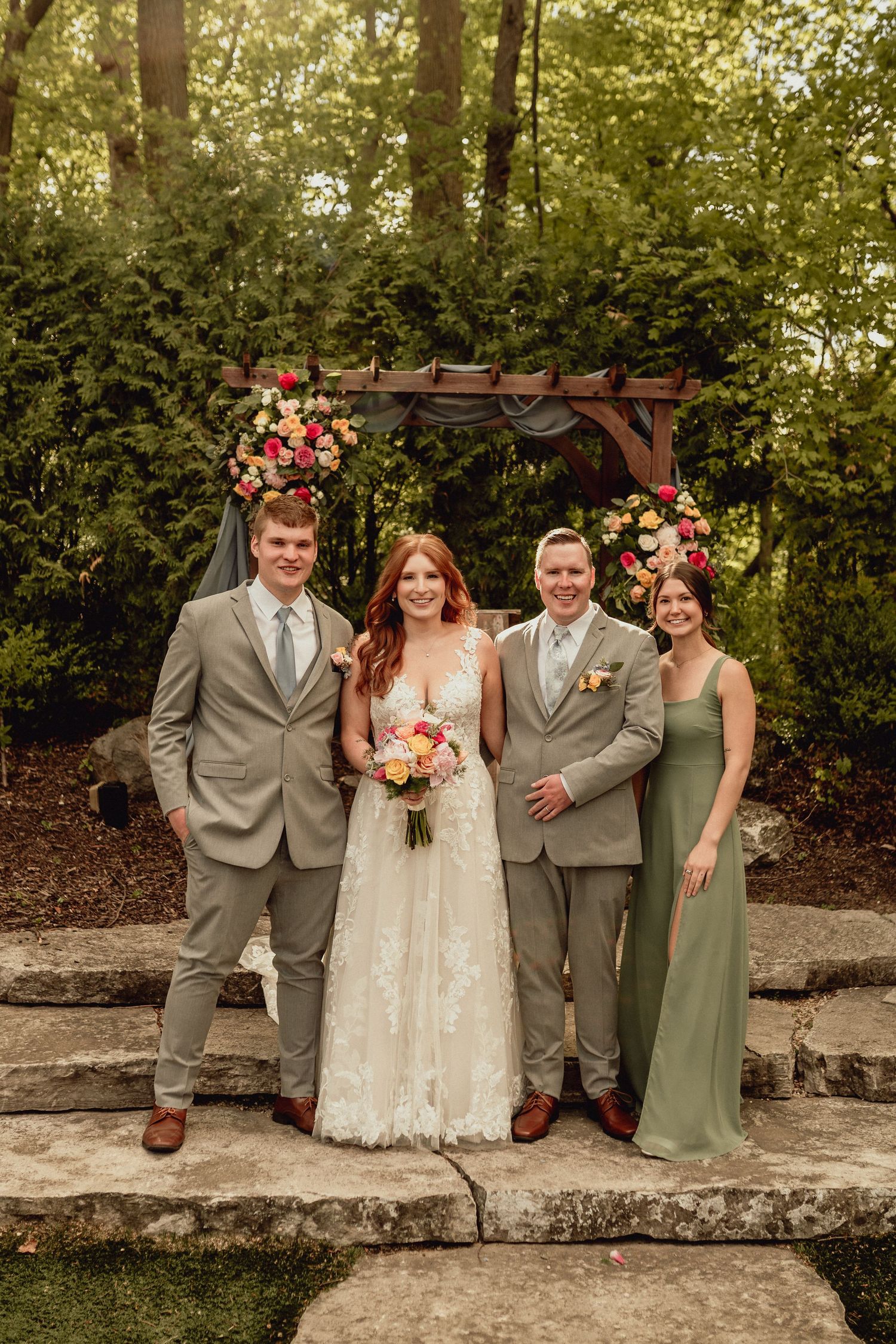 Wedding party poses under floral arch on stone steps in garden setting with greenery backdrop.