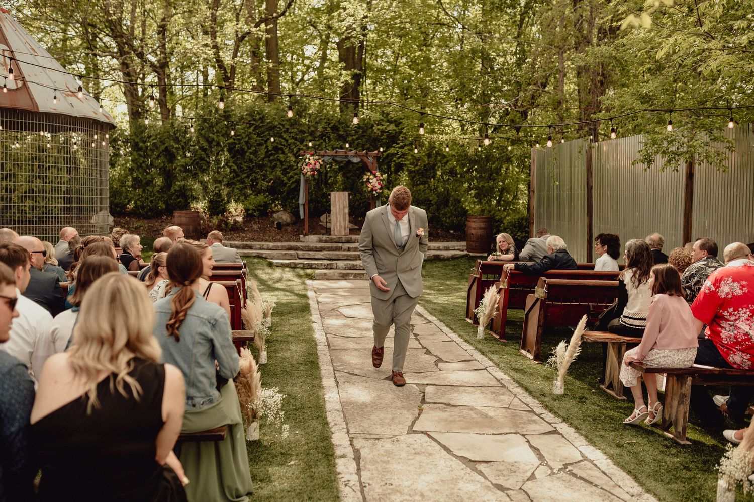 A joyous outdoor wedding ceremony takes place in a garden courtyard with stone pathways and green trees.