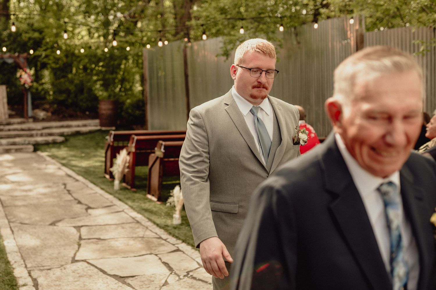 A couple in formal wedding attire walks through a garden setting during an outdoor ceremony.
