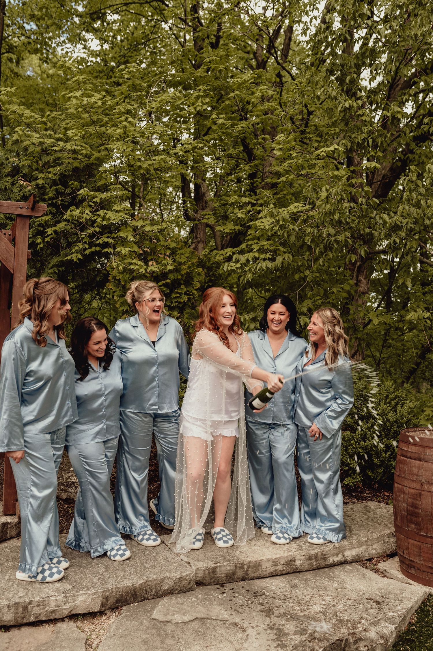 A wedding party in matching light blue satin outfits poses playfully on a stone wall surrounded by green trees.