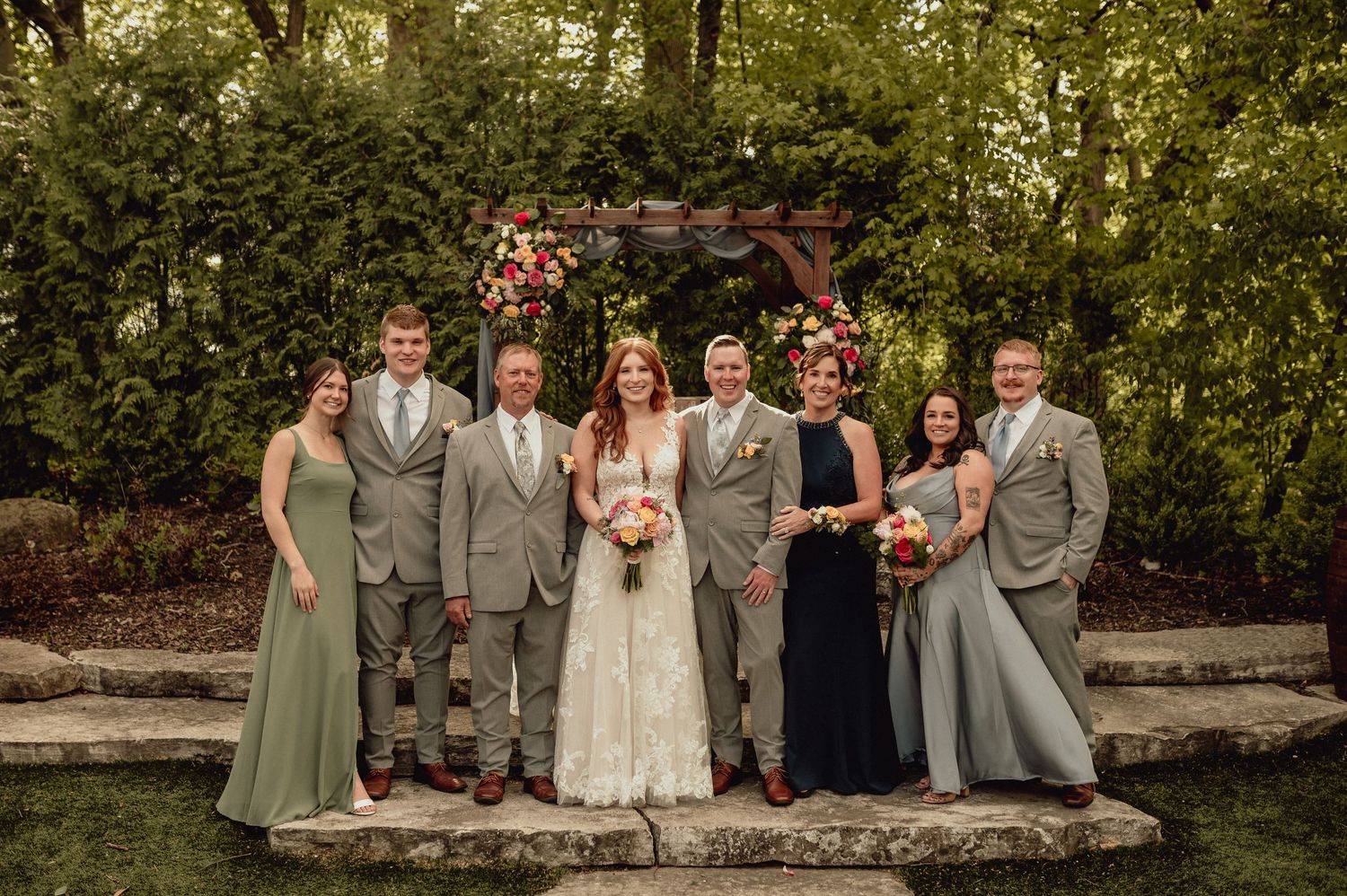 Wedding party in sage green dresses and gray suits poses outdoors on stone wall during golden hour.