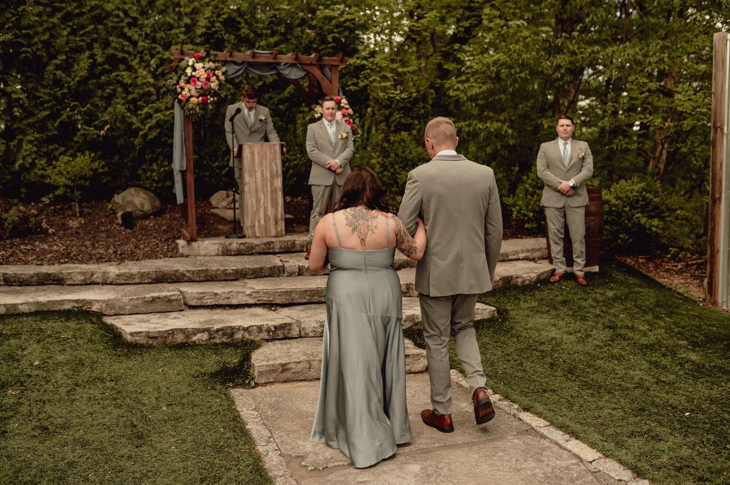 Wedding party members walk down stone steps in an outdoor garden setting wearing gray and tan formal attire.
