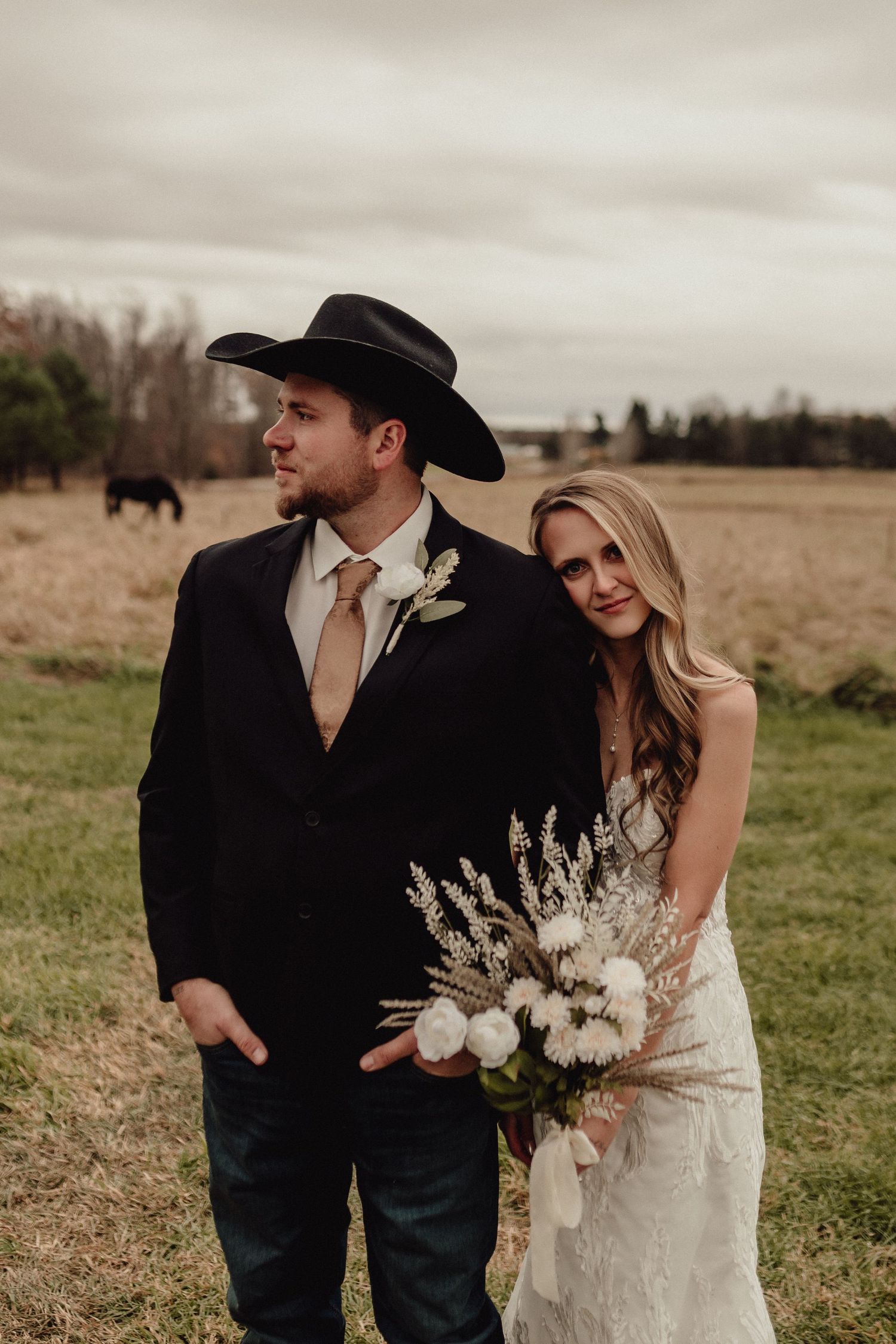 A couple in western wedding attire embraces in a rustic field setting with dried grass and moody sky.