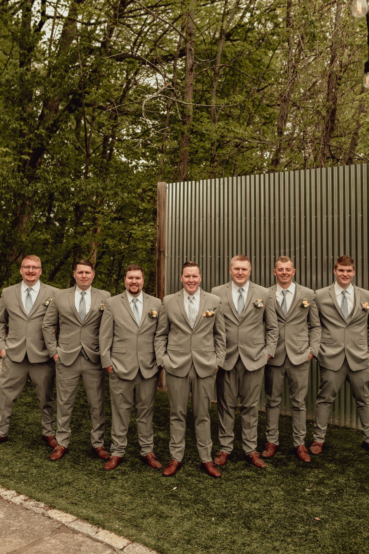 A group in matching light gray suits and brown shoes stand in a line against a metal wall backdrop near trees.