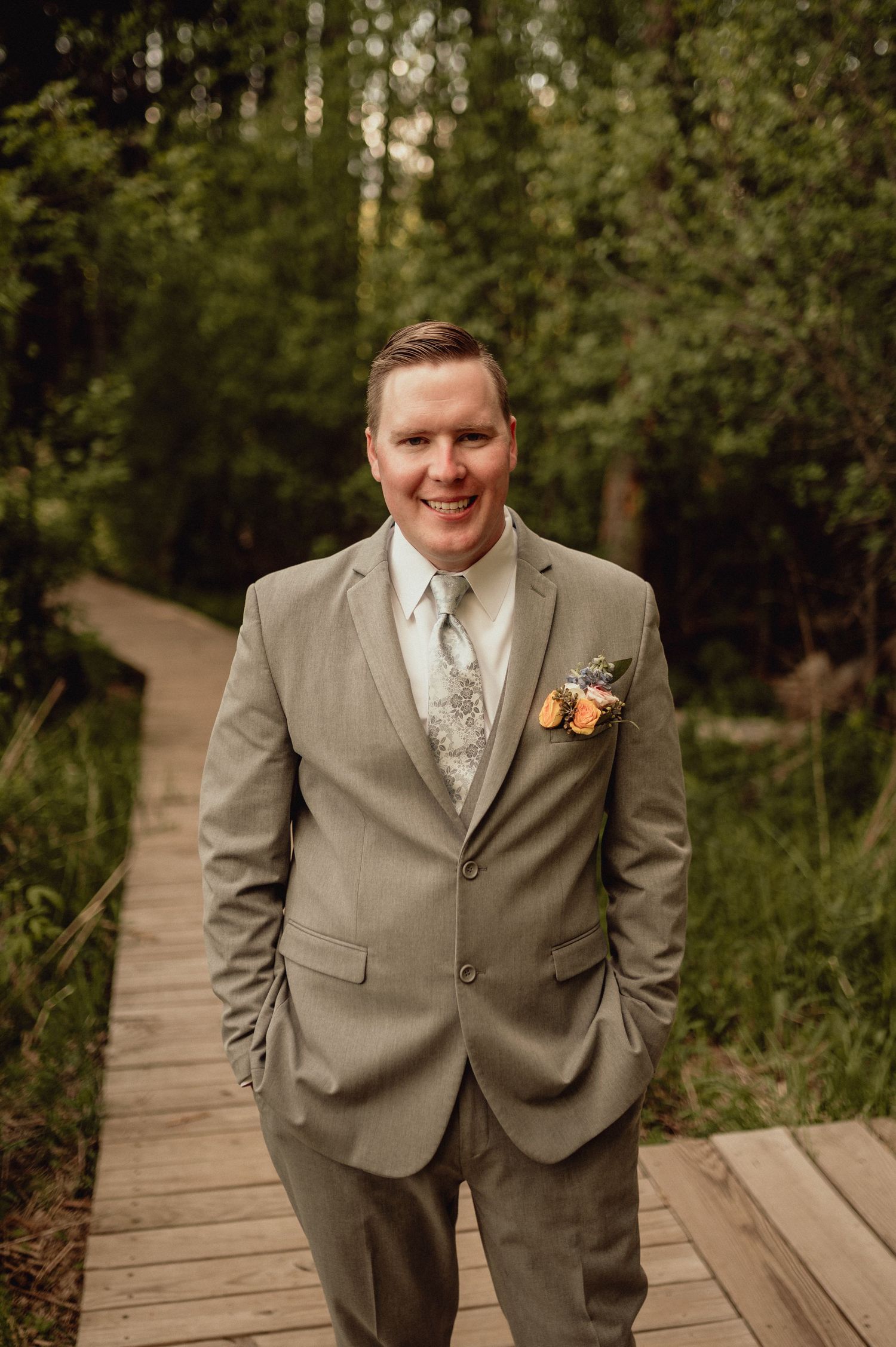 A groom in a light gray suit and floral boutonniere smiles while standing on a wooden boardwalk in a forest setting.