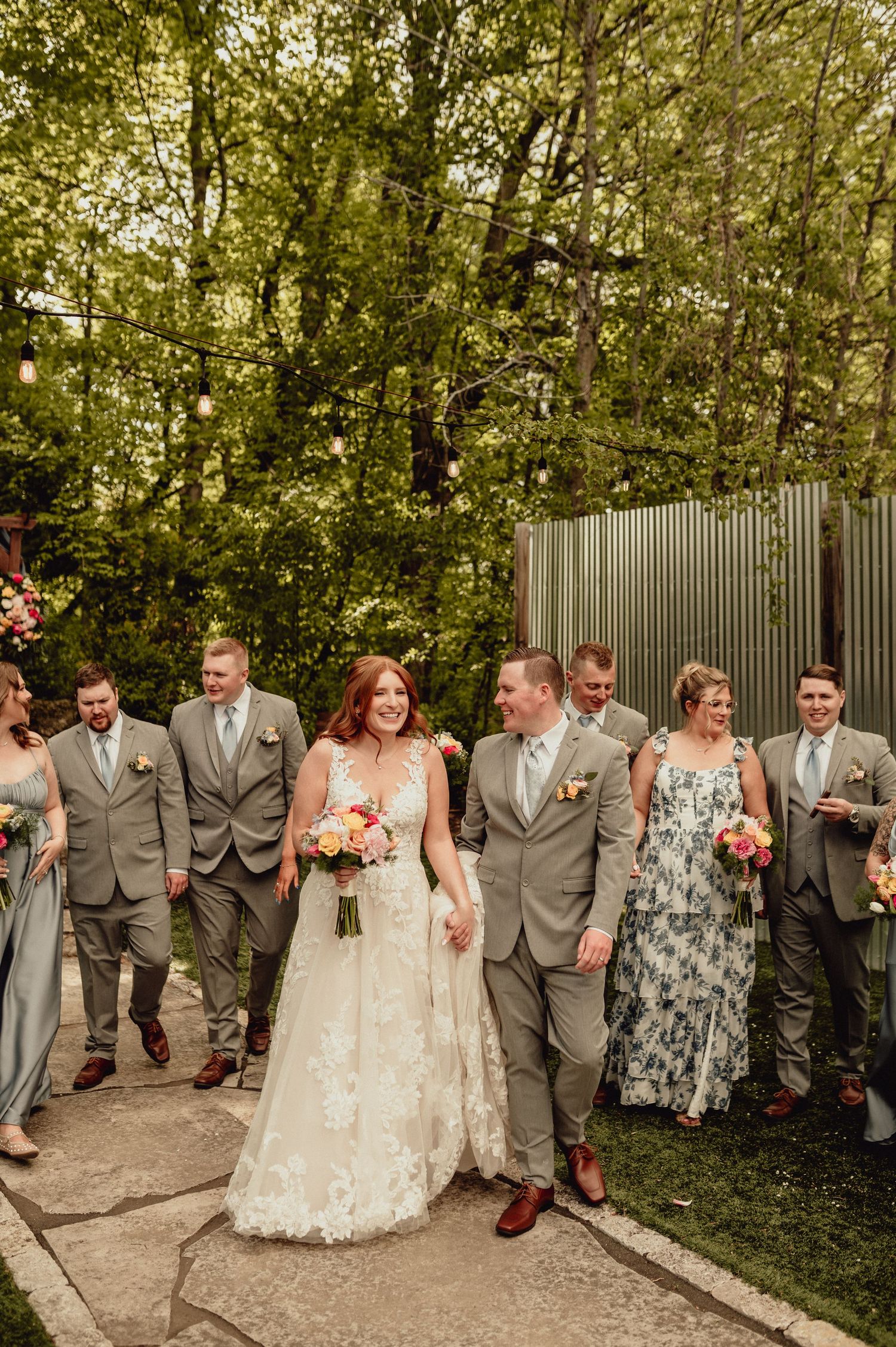 Wedding party in gray suits and floral dresses poses together in a garden setting with greenery backdrop.