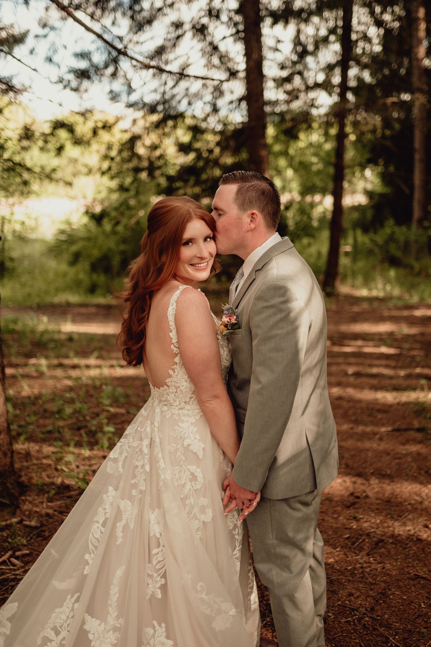 A romantic wedding couple shares intimate moments in a sunlit forest setting with pine needles covering the ground.