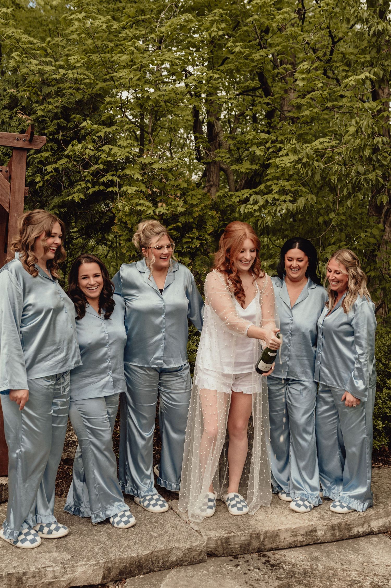 Bridesmaids in matching light blue satin pajamas pose together on a stone wall outdoors.