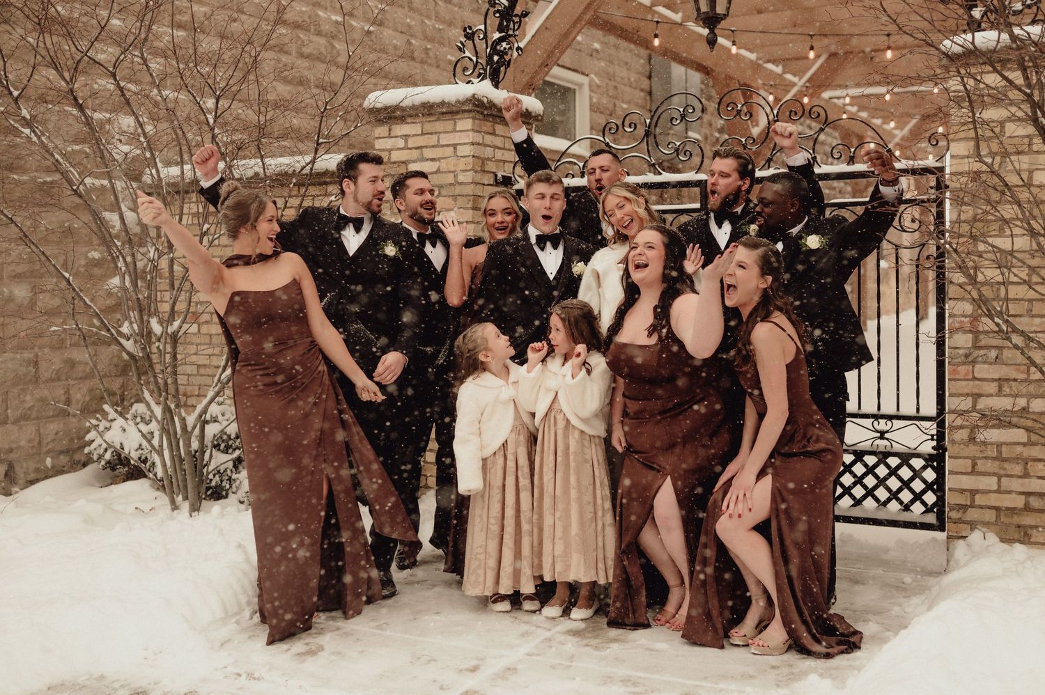 Wedding party in brown and beige attire poses in front of brick wall during winter snowfall.