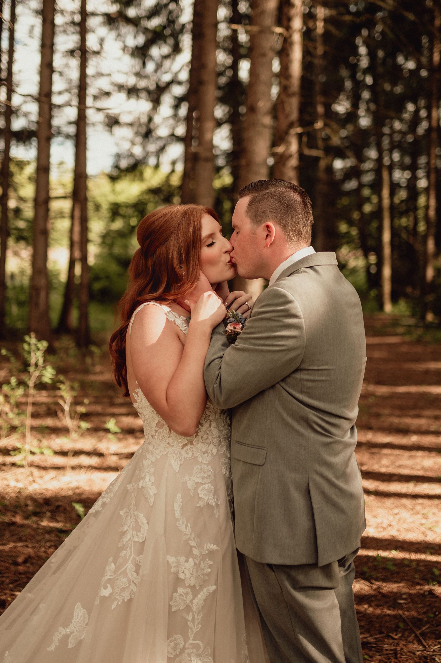 A couple shares a romantic kiss in a forest setting with sunlight filtering through pine trees.