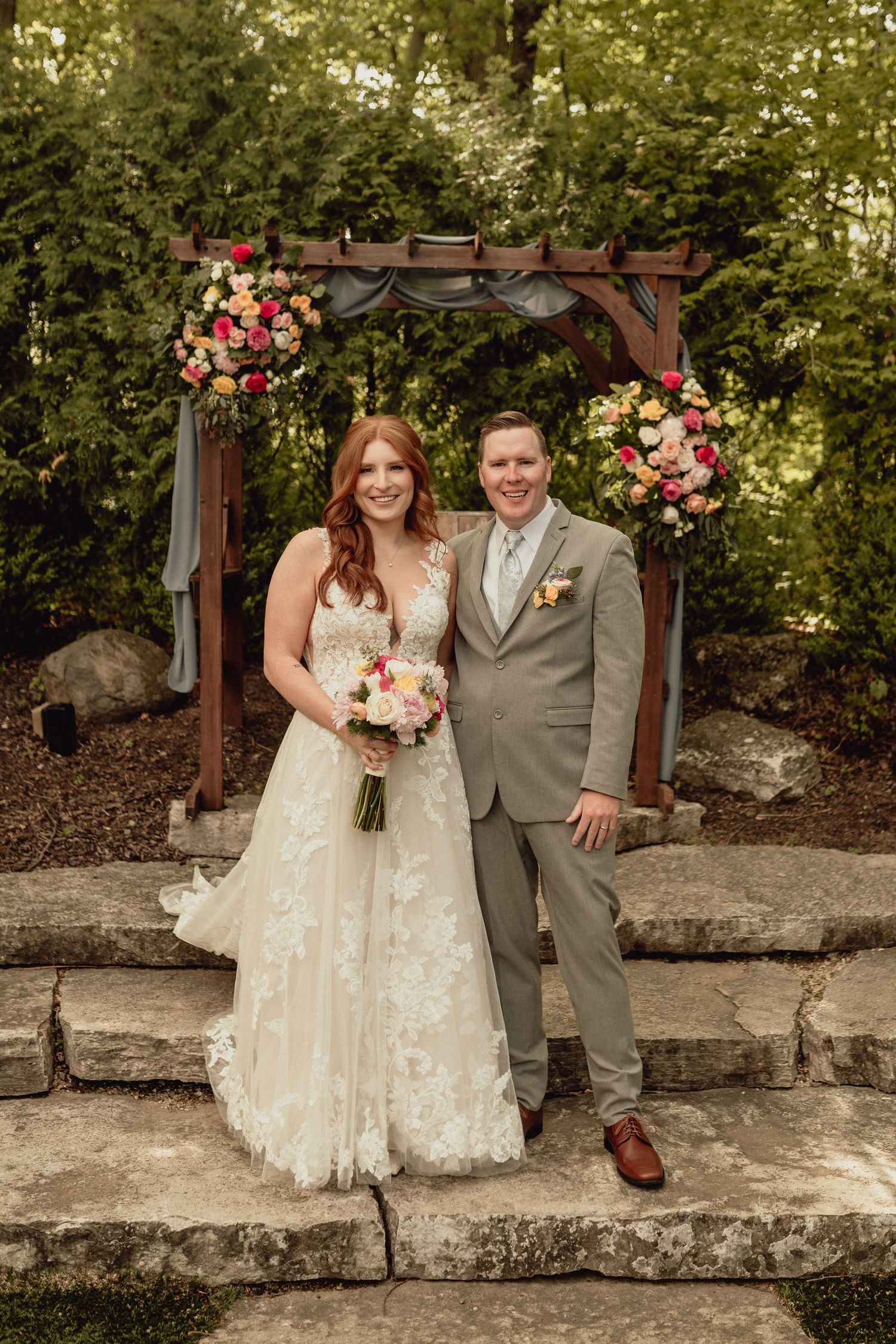 A couple in wedding attire sits on stone steps with floral decorations in an outdoor garden setting.