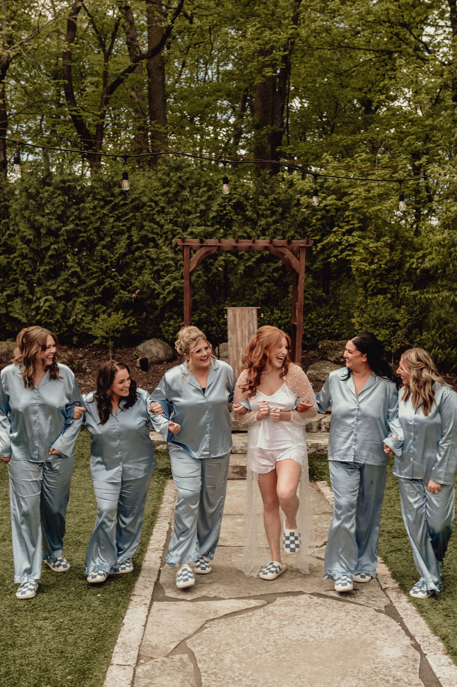 A group in matching blue satin pajamas walks together on a garden path while sharing laughs and smiles.