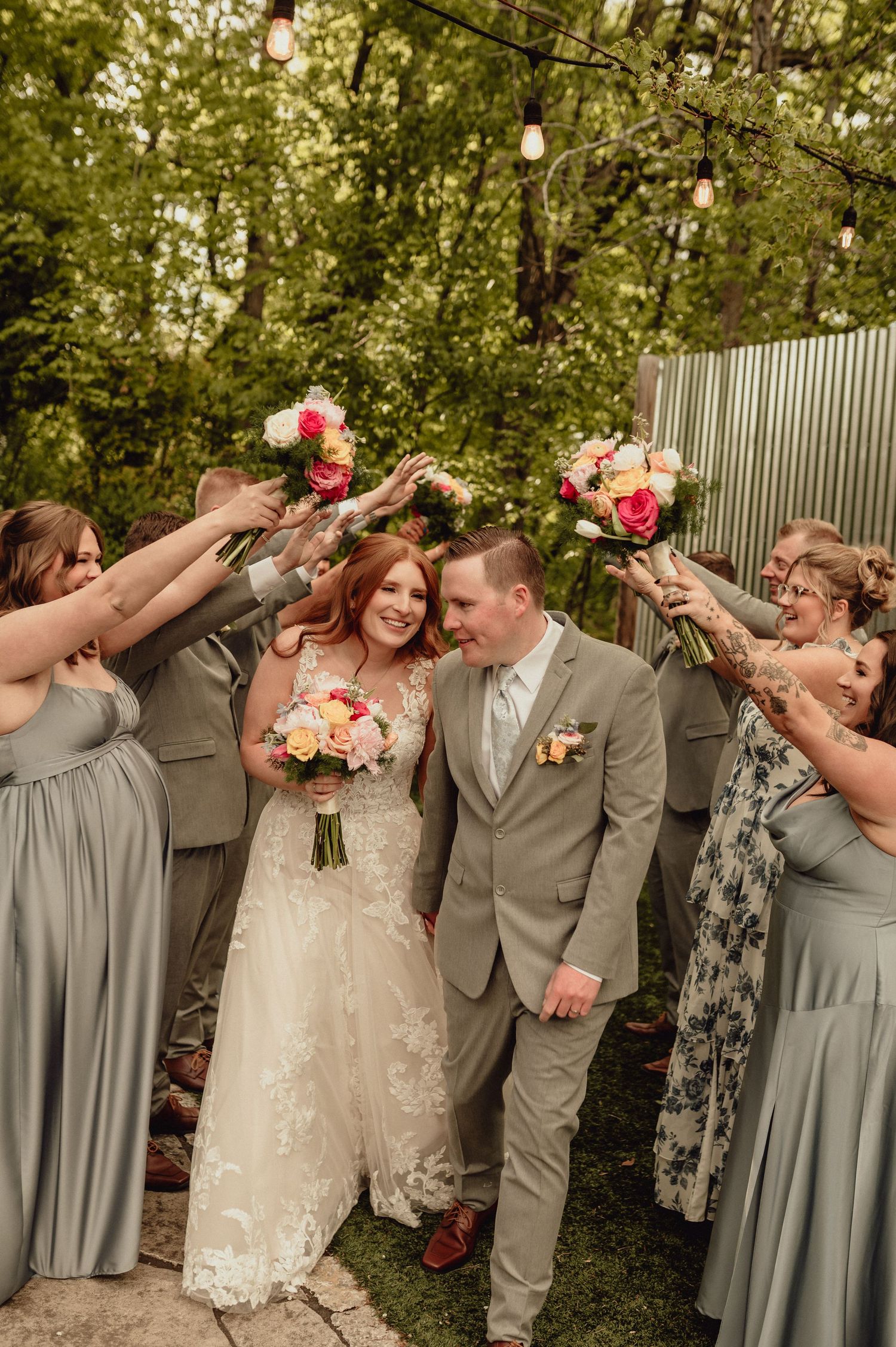 Wedding party in gray attire poses playfully together in a garden setting with pink floral bouquets.