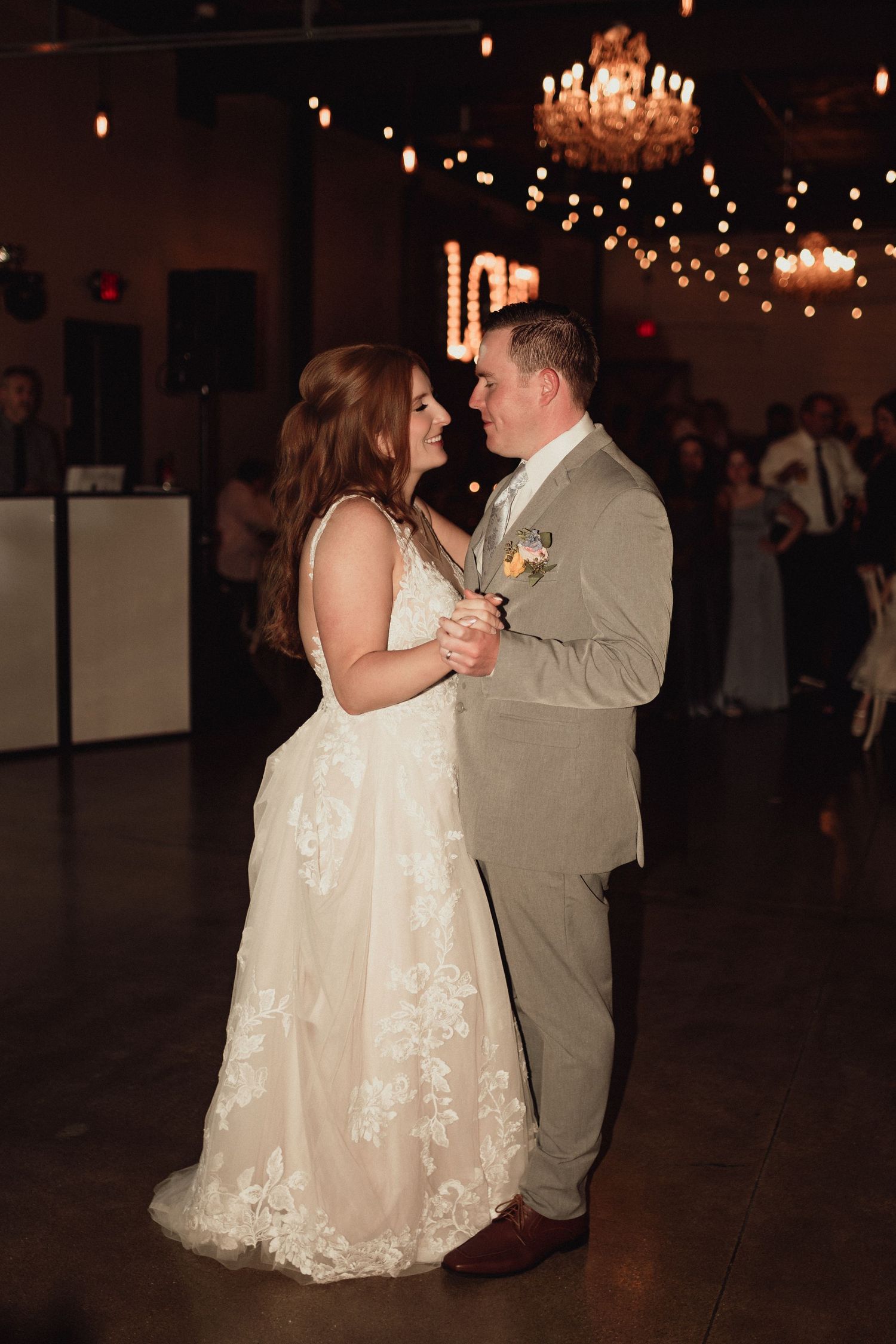 A couple shares their first dance at a wedding reception under a crystal chandelier and string lights.