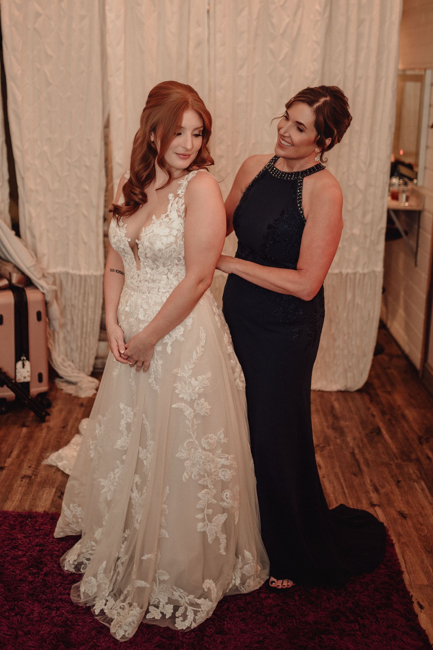 A bride in a lace wedding gown stands with a woman in a black formal dress on hardwood floors during preparations.
