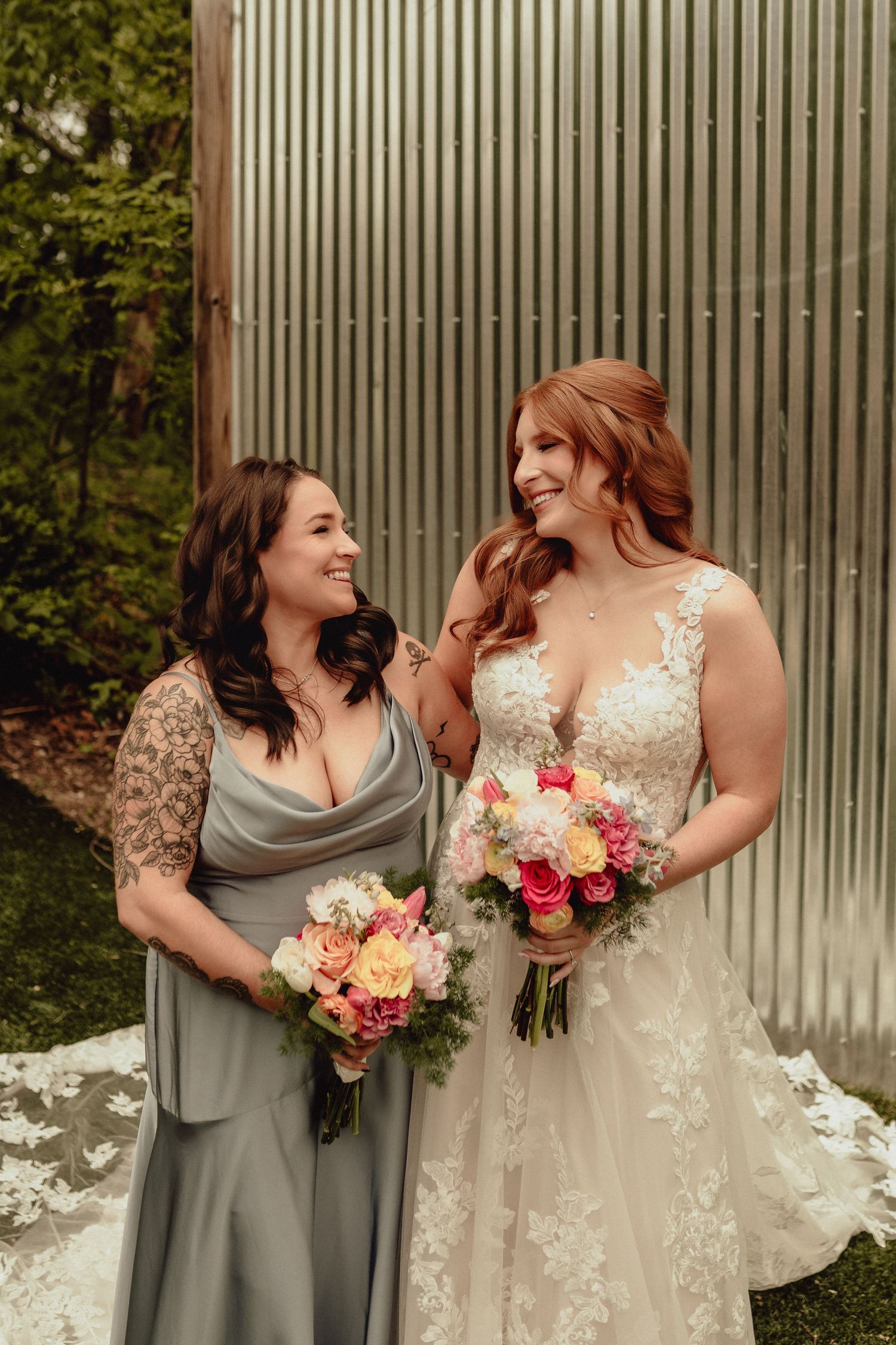 Two people in wedding attire share candid laughing moments while holding floral bouquets against a metal wall backdrop.