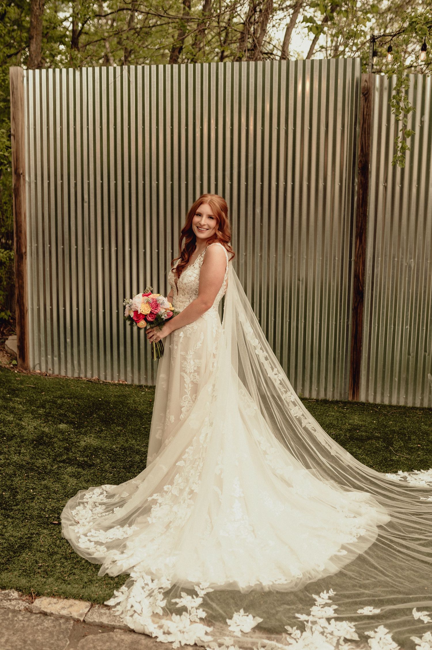 A bride in a flowing lace wedding gown with a dramatic cathedral veil poses in a garden setting.