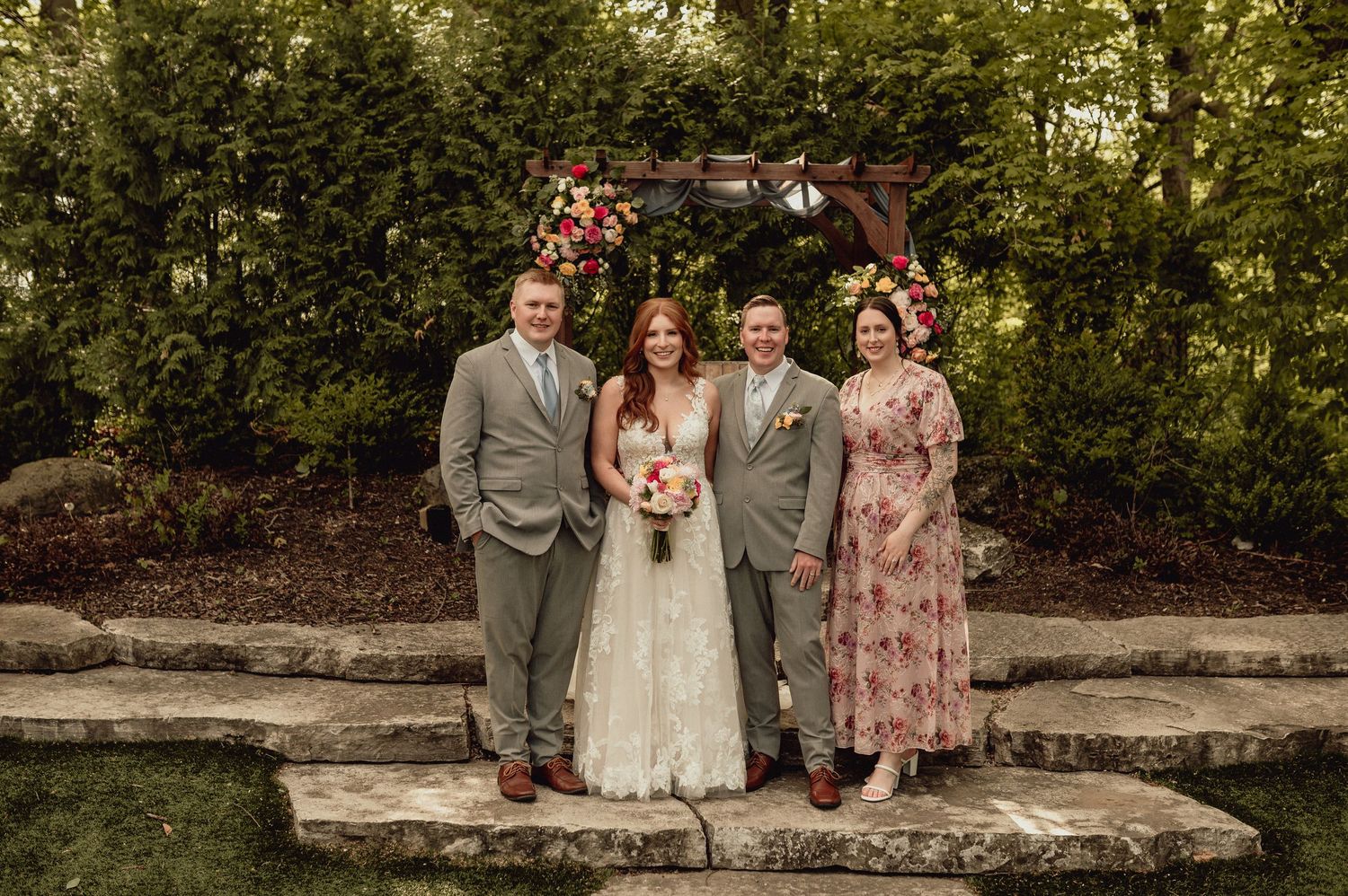 A small wedding party stands together on stone steps in front of a lush garden backdrop during an outdoor ceremony.