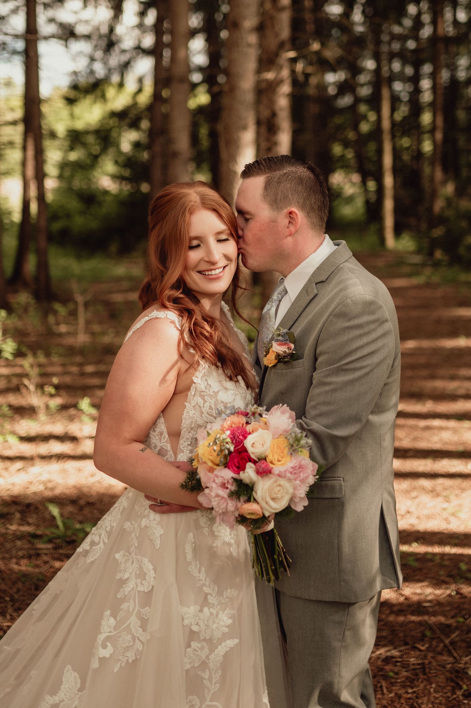 A couple in wedding attire shares romantic moments in a sunlit forest setting with autumn leaves on the ground.