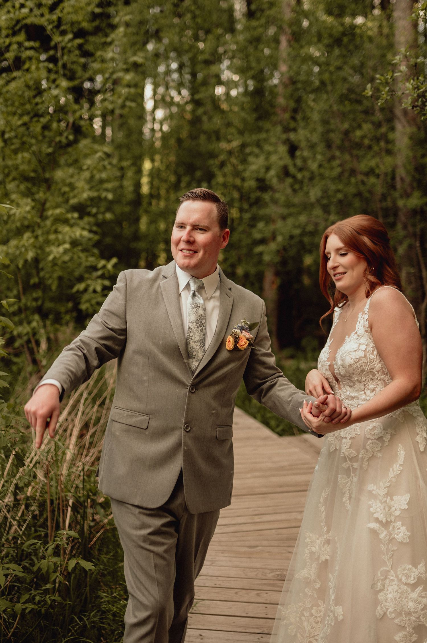 A joyful couple in wedding attire share candid laughing moments on a wooden boardwalk surrounded by green forest.