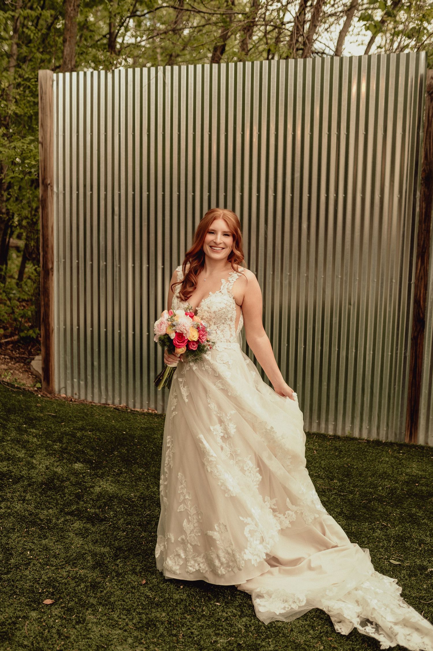 A bride in a lace wedding gown poses outdoors against a corrugated metal wall in a garden setting.