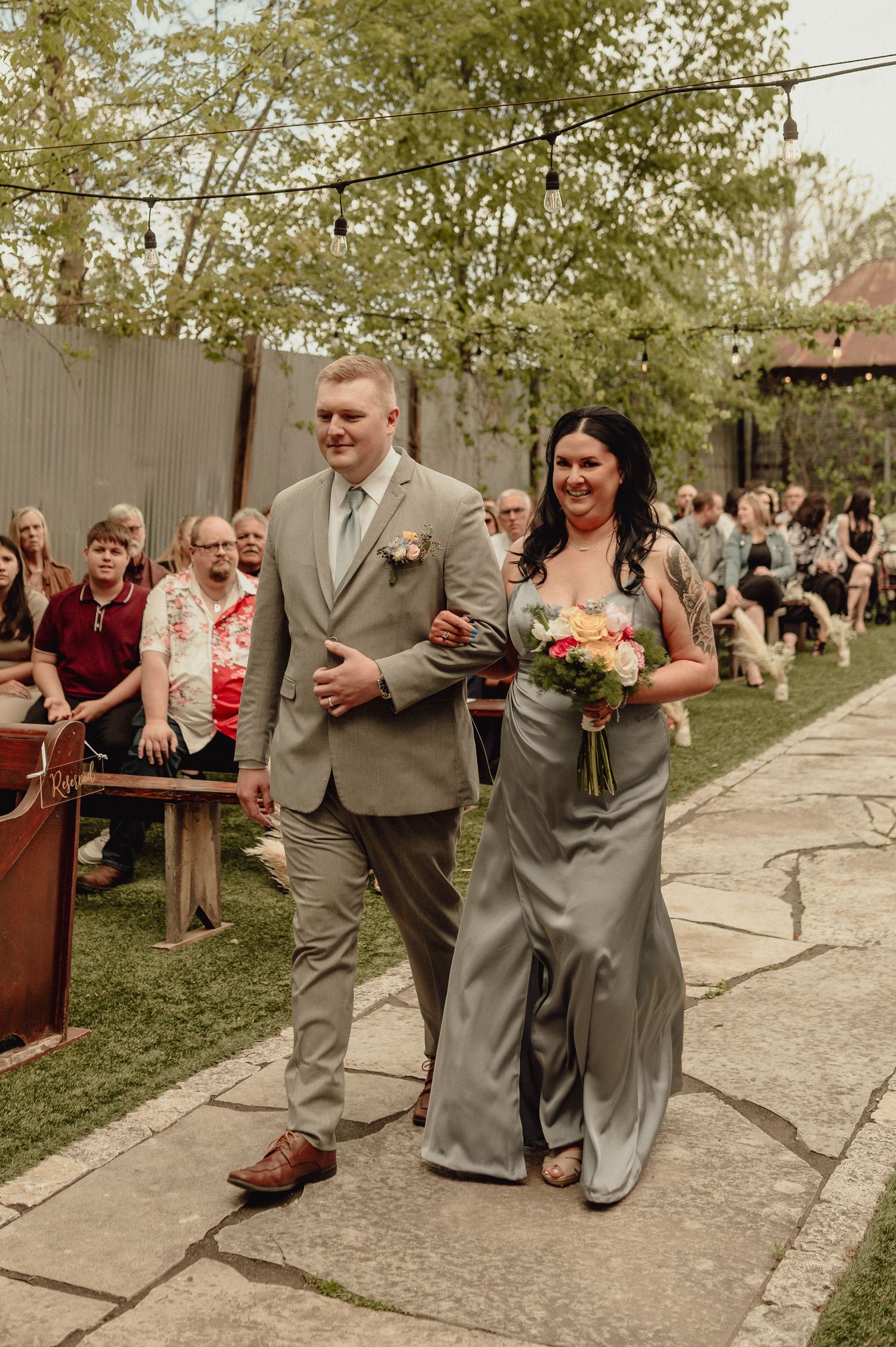 Wedding party members in gray attire walk down stone pathway during outdoor ceremony as guests look on.