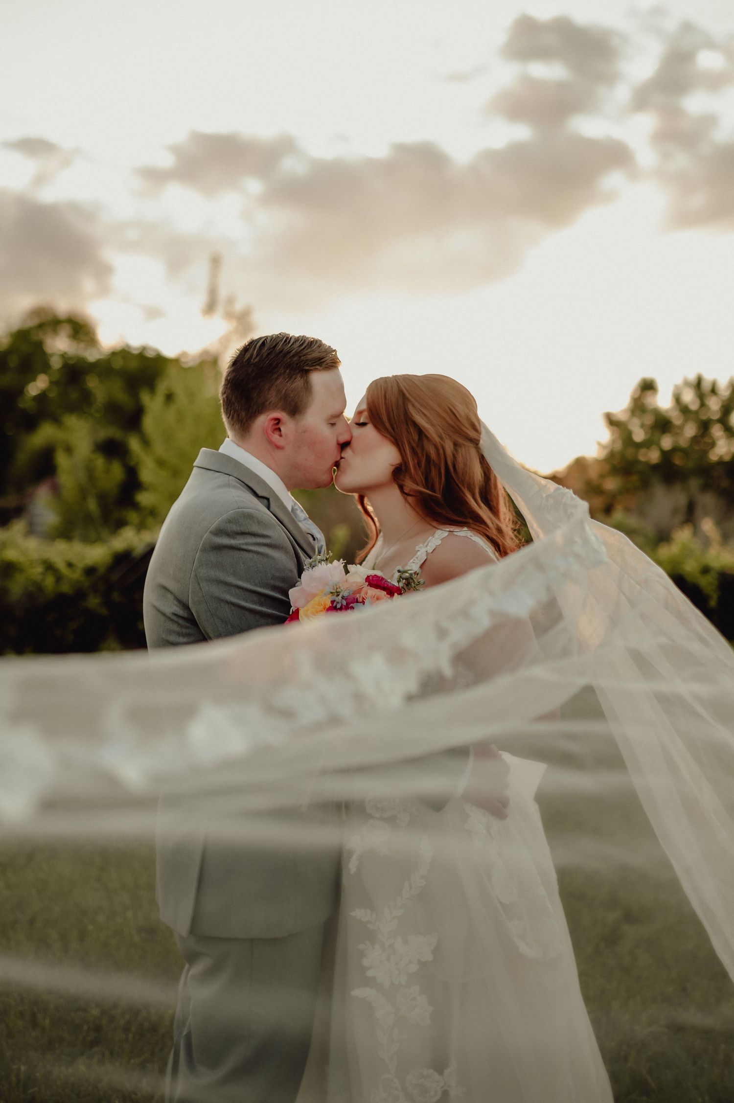 A romantic wedding couple shares a kiss at sunset while the bride's veil flows gracefully in the wind.
