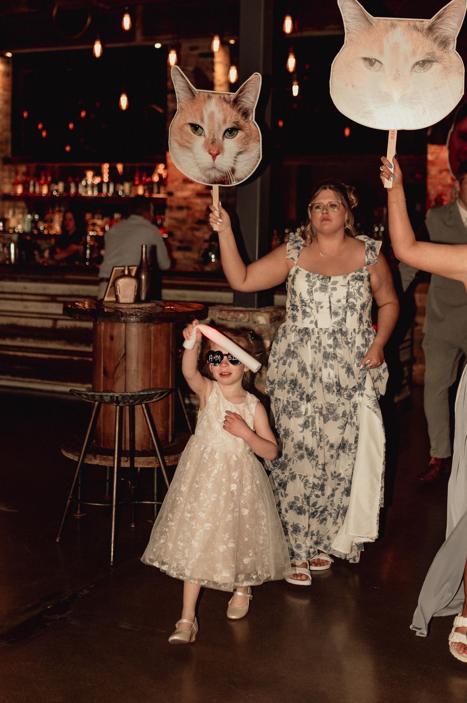 People at a celebration holding large cat face cutouts while dancing in a dimly lit venue with hanging lights.