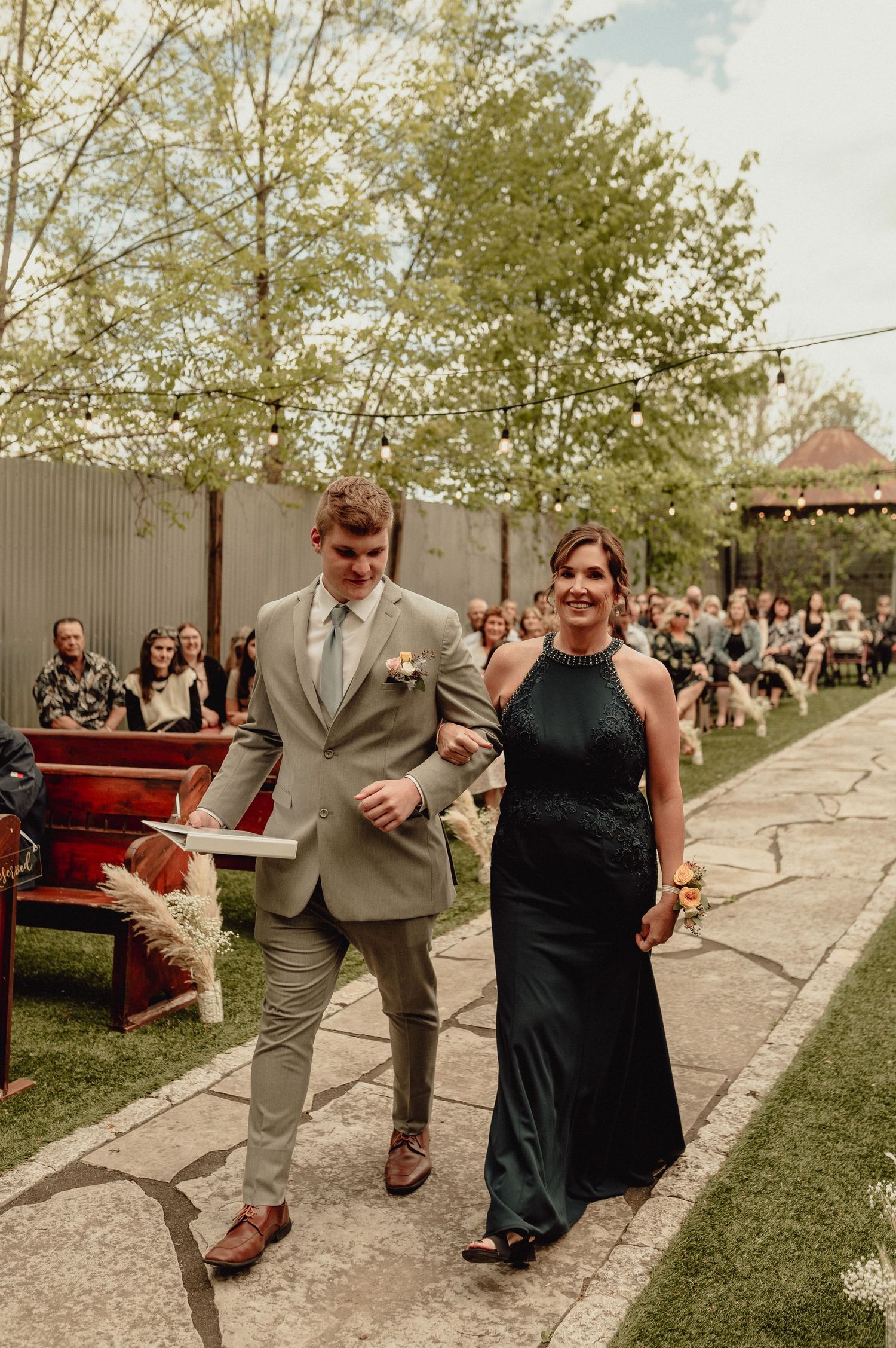 A couple in formal attire runs playfully down a path at an outdoor event with guests visible in the background.