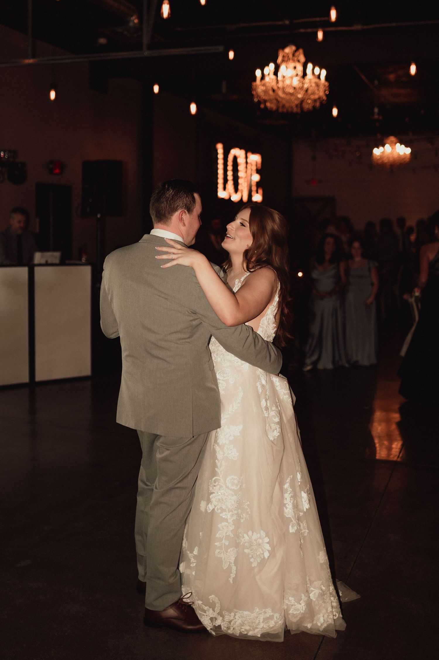 A couple shares an intimate dance moment at a dimly lit wedding reception with chandeliers overhead.