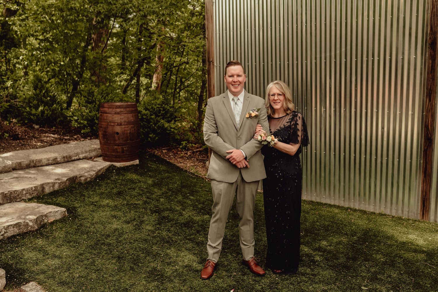 A couple stands together in a garden setting with corrugated metal wall and landscaping in the background.