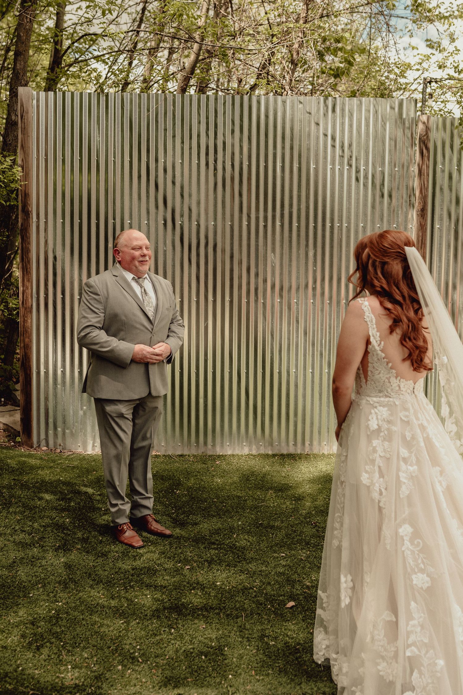 A touching wedding moment captured outdoors with a lace wedding dress and grey suit against a corrugated metal backdrop.