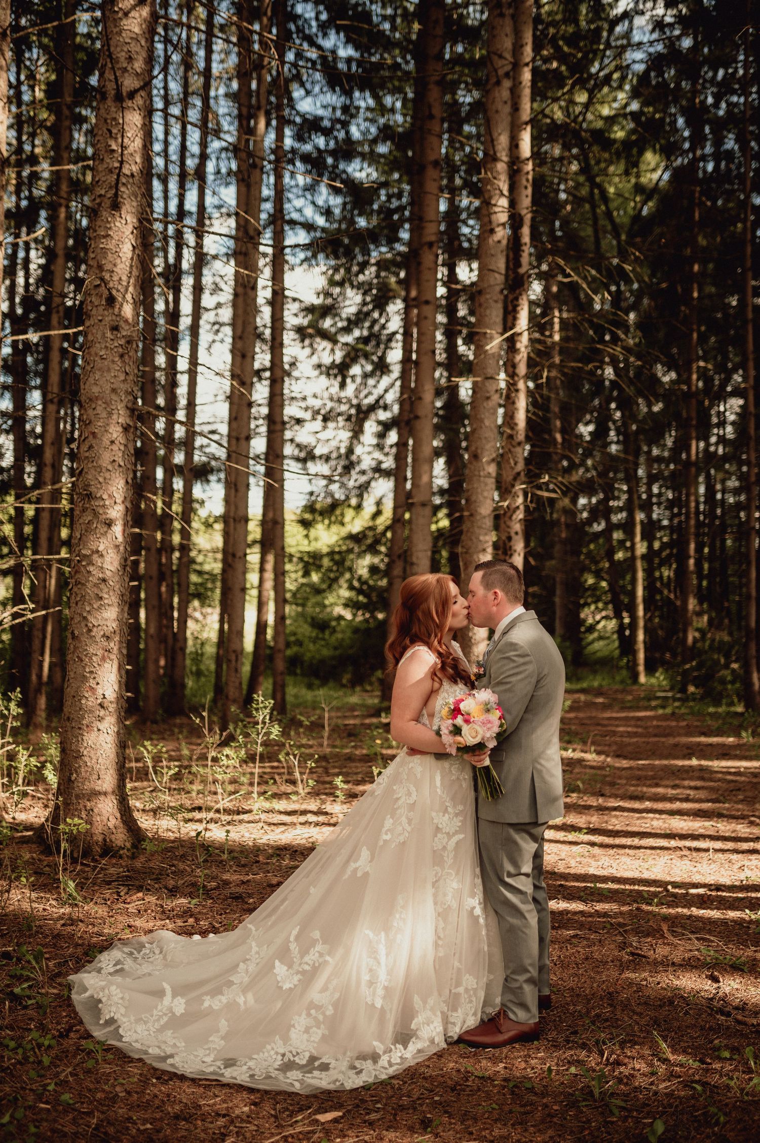 A romantic wedding couple embraces in a sunlit forest setting with tall pine trees creating a natural backdrop.
