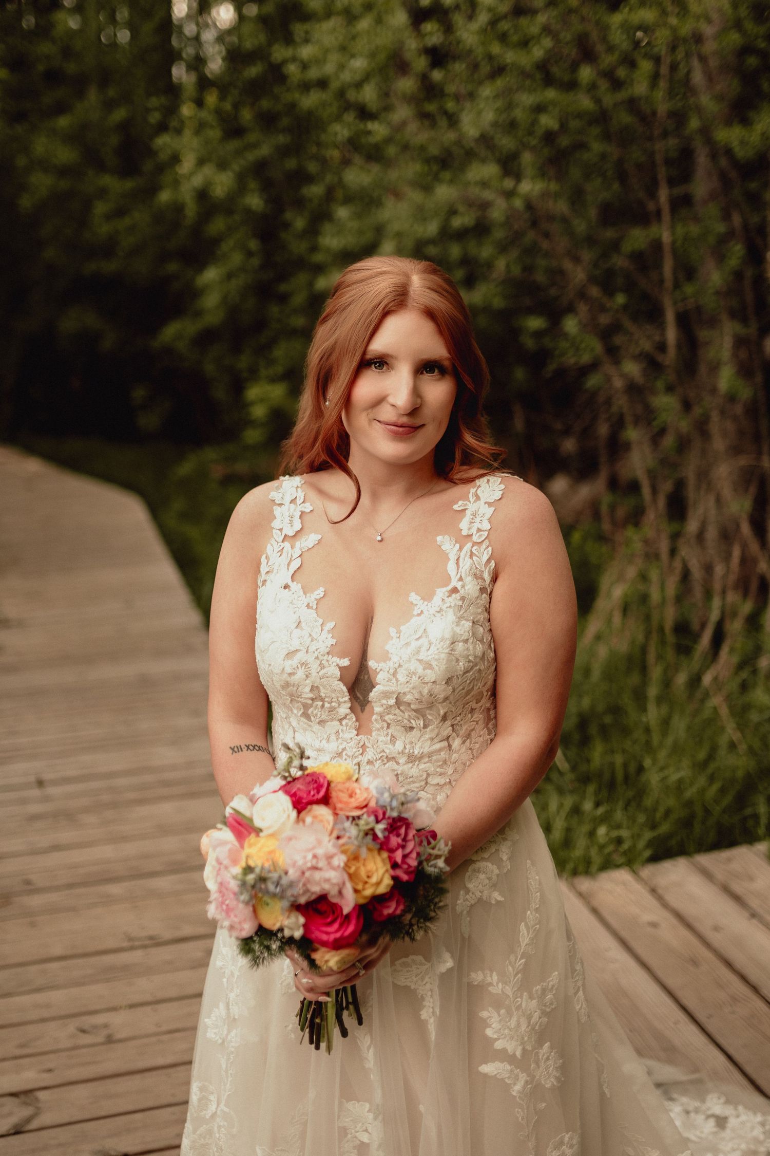 A bride in a lace wedding dress poses with a colorful bouquet on a wooden boardwalk surrounded by greenery.