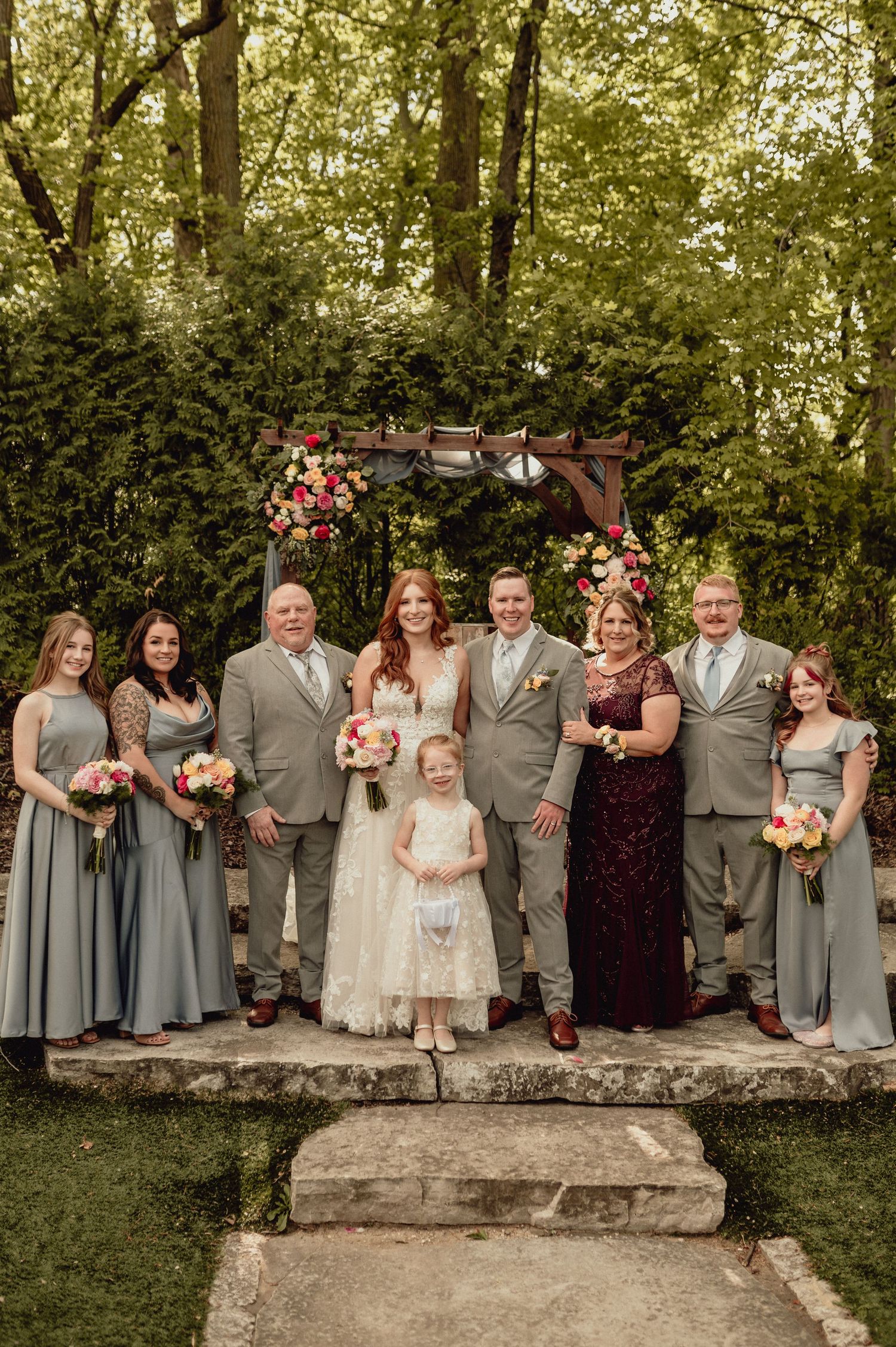Wedding party in sage green and burgundy attire poses together under floral arch in outdoor garden setting.
